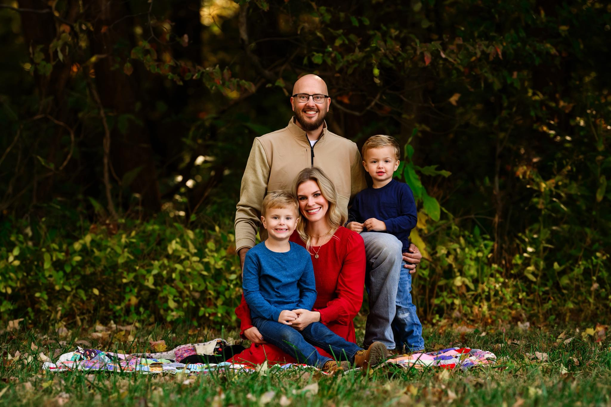 A family of four posing outdoors in front of trees. The mother and two young boys are seated on a blanket on the ground, with the father standing behind them. All are smiling and dressed casually, with autumn-colored foliage in the background.