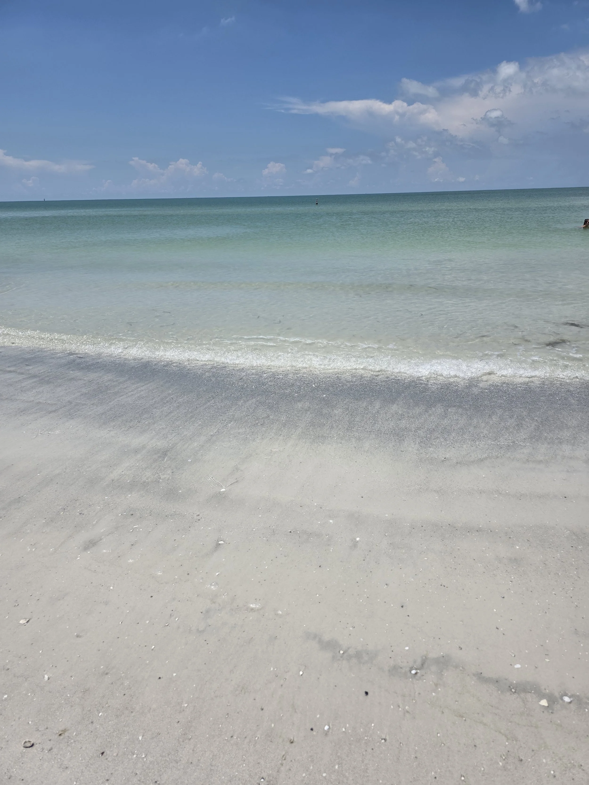 Calm ocean waves gently crash on a pristine sandy beach with clear blue sky and scattered clouds.