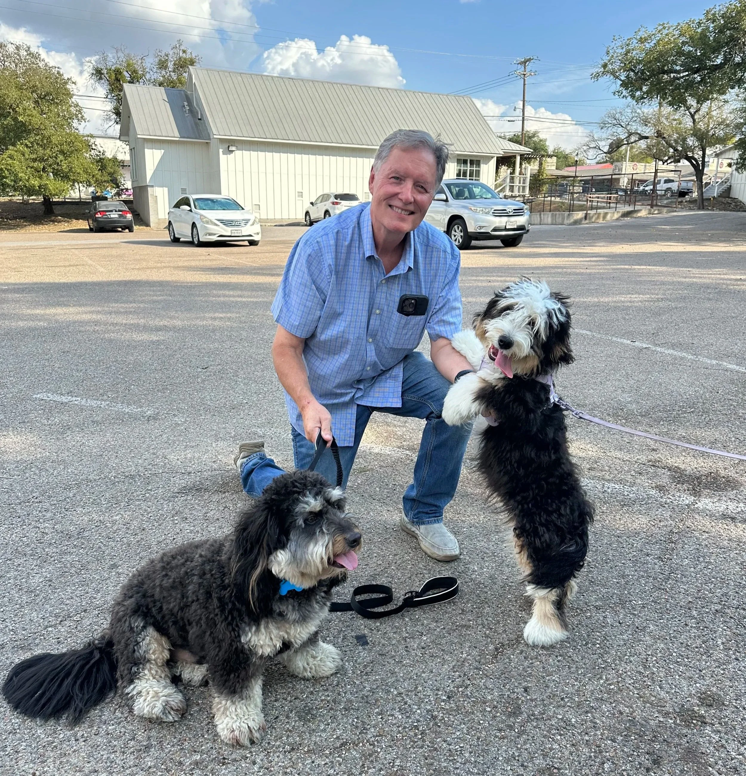A man in a blue shirt smiling and kneeling on the pavement with two black and white dogs, one sitting in front and one standing beside him. There are cars parked in the background and a white building with a gray roof. The sky is partly cloudy.