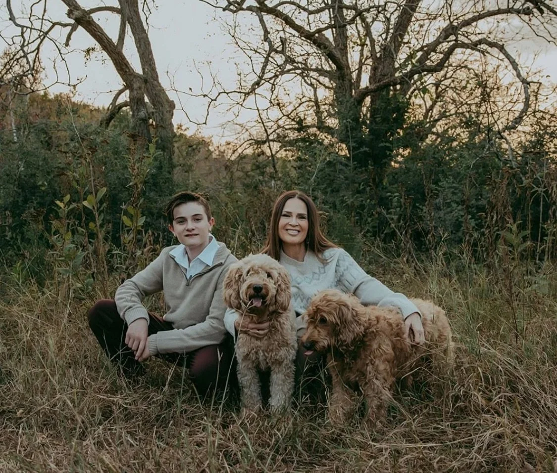 A woman and a teenage boy sitting in a field with two dogs, surrounded by tall grass and trees, during sunset.