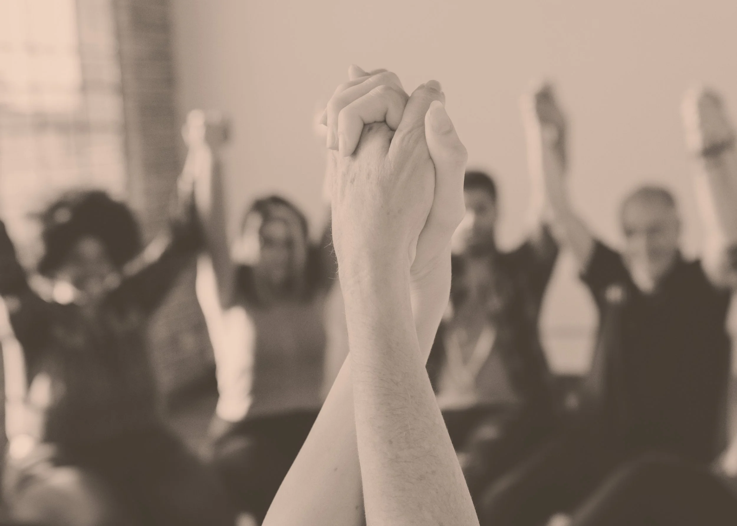 Close-up of two hands clasped together in prayer or support, with a group of people in the background raising their hands, in a monochrome setting.