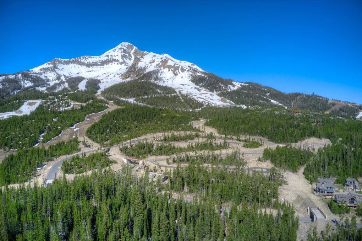 Snow-capped mountain with winding roads and dense green forest at its base under a clear blue sky.