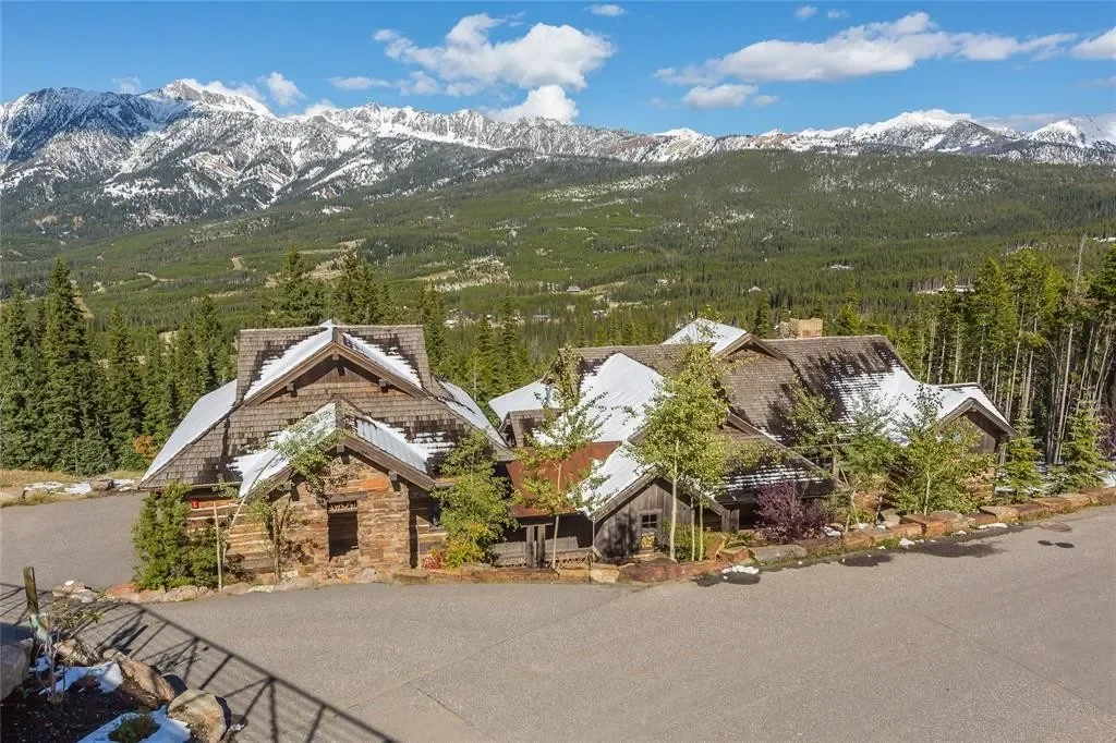 A mountain cabin surrounded by trees, with snow-capped mountains in the background and a gravel driveway in the foreground.