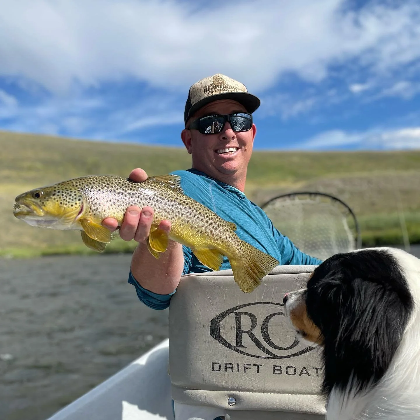 A man in a blue shirt and sunglasses holding a large rainbow trout fish on a boat with a black and white dog beside him, with a background of hilly landscape and a partly cloudy sky.