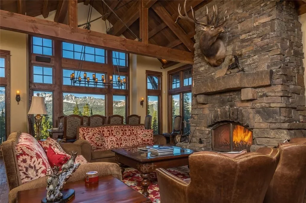 Living room with large stone fireplace topped with a mounted deer head, surrounded by wooden beams and large windows showing mountain views, and furnished with patterned couches and leather armchairs.