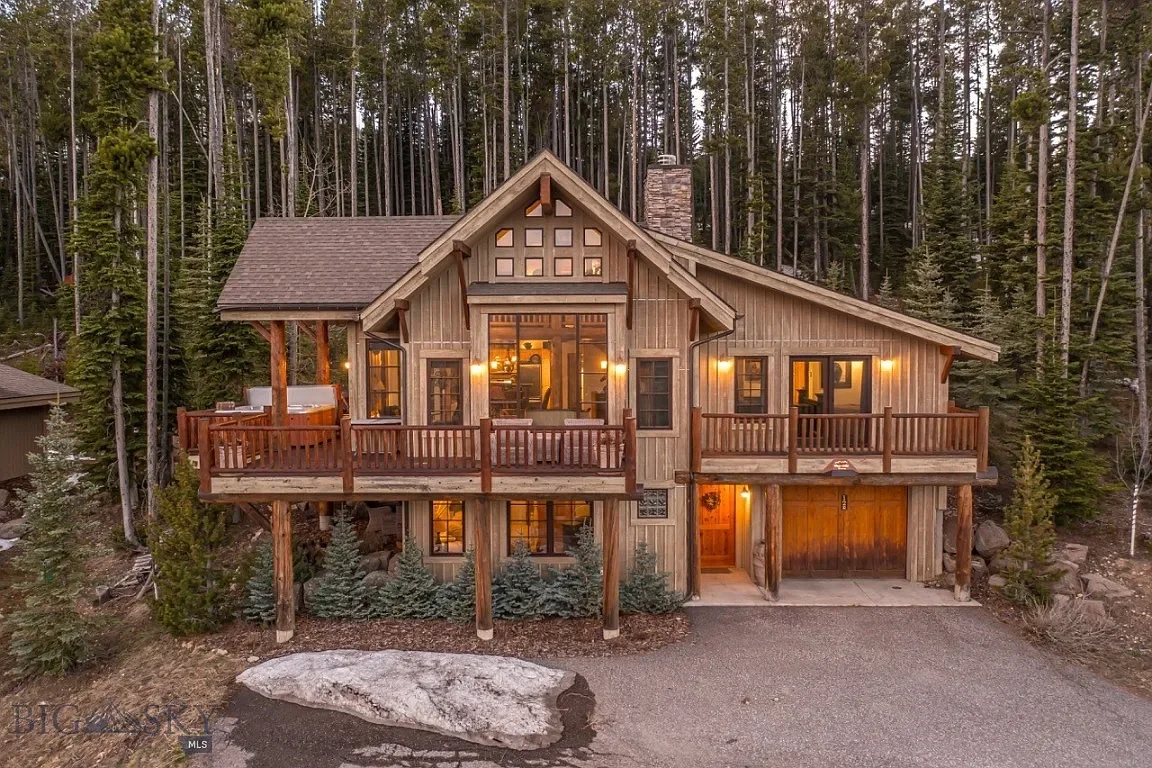 A rustic two-story house surrounded by tall trees with a gravel driveway and outdoor lighting.
