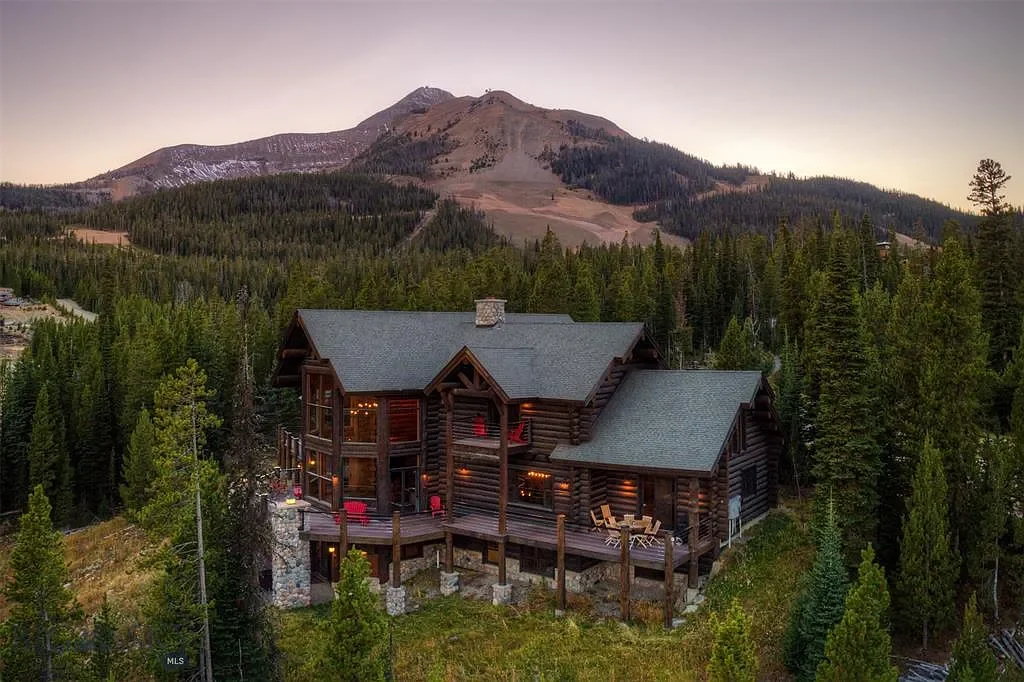 A large rustic log cabin with a deck, surrounded by pine trees, with a mountain in the background at dusk.