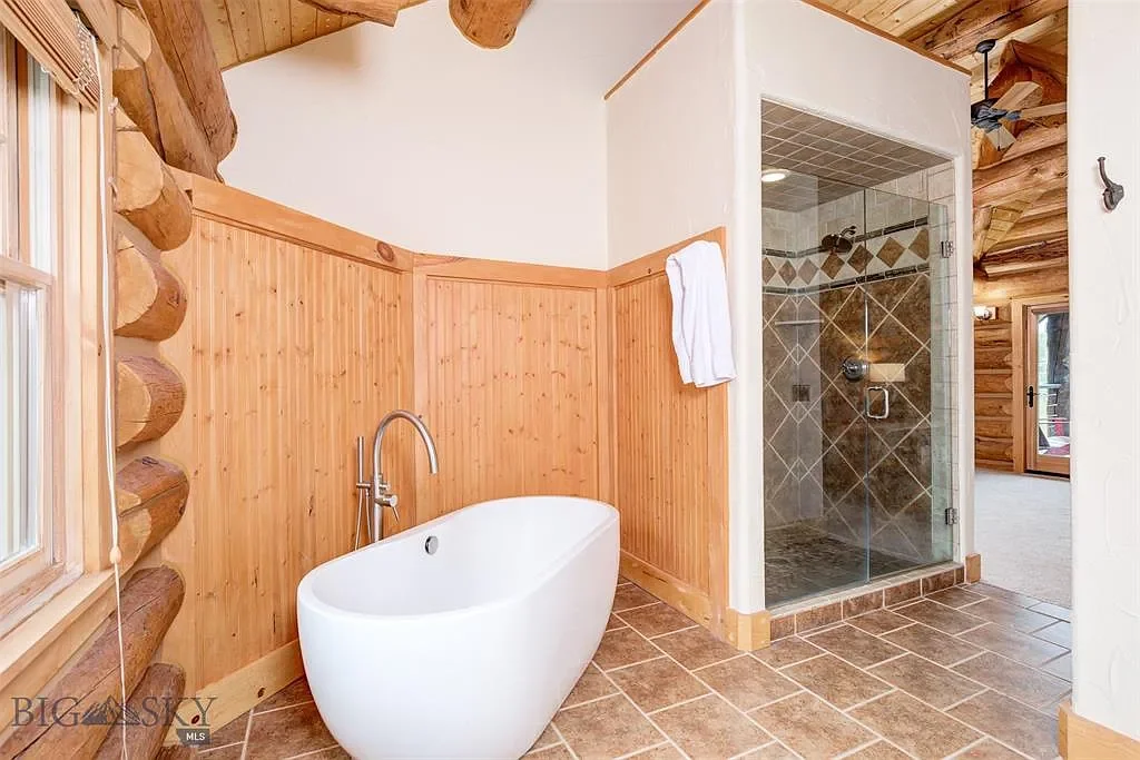 Bathroom with wood-paneled walls, a white freestanding bathtub, a glass-enclosed shower, and a window with logs and ceiling in on the right side.