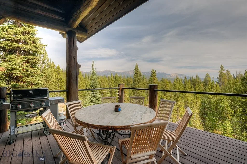 A wooden deck with a round table surrounded by six wooden chairs, overlooking a forested landscape with mountains in the distance under a partly cloudy sky.
