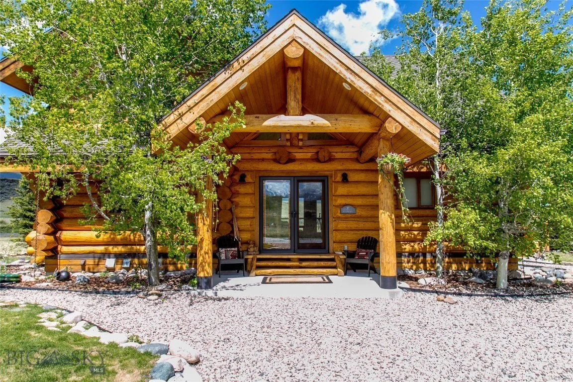 Front view of a log cabin named Big Sky. It has a wooden porch with steps, two black chairs, and hanging flower baskets. There are trees and a gravel pathway in front, under a partly cloudy blue sky.
