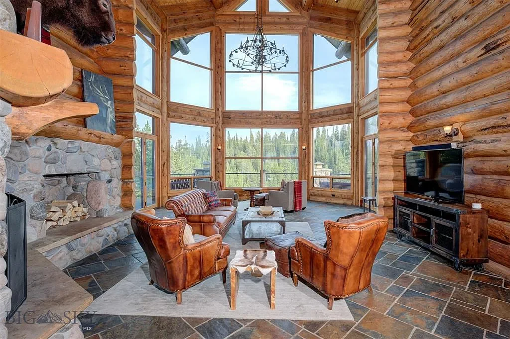 A spacious living room with large windows, wooden walls, a stone fireplace, leather and fabric furniture, and a mountain view outside.