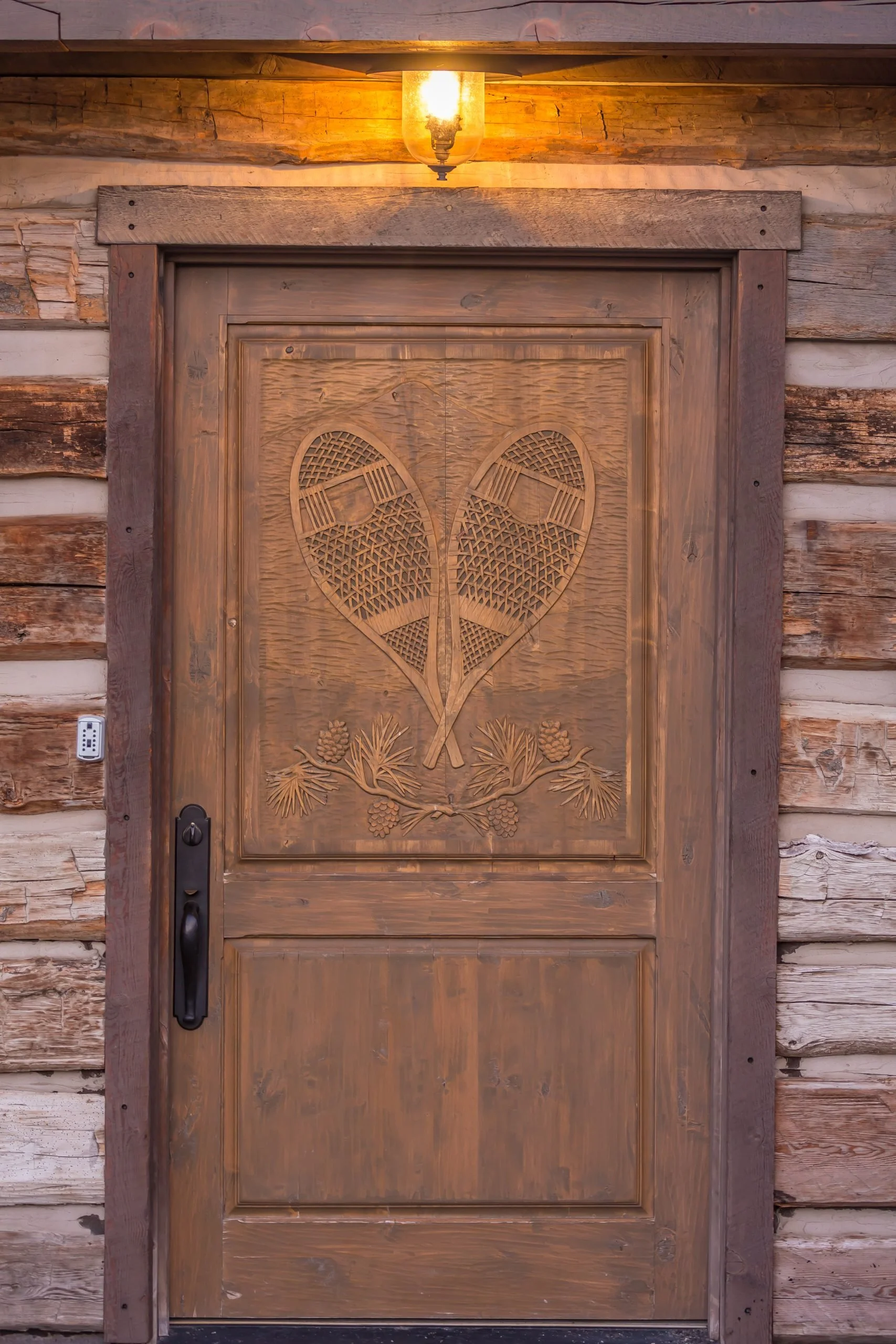 Wooden door with a carved heart-shaped design of two tennis rackets and pinecones, set in a rustic log cabin wall illuminated by a warm outdoor light.