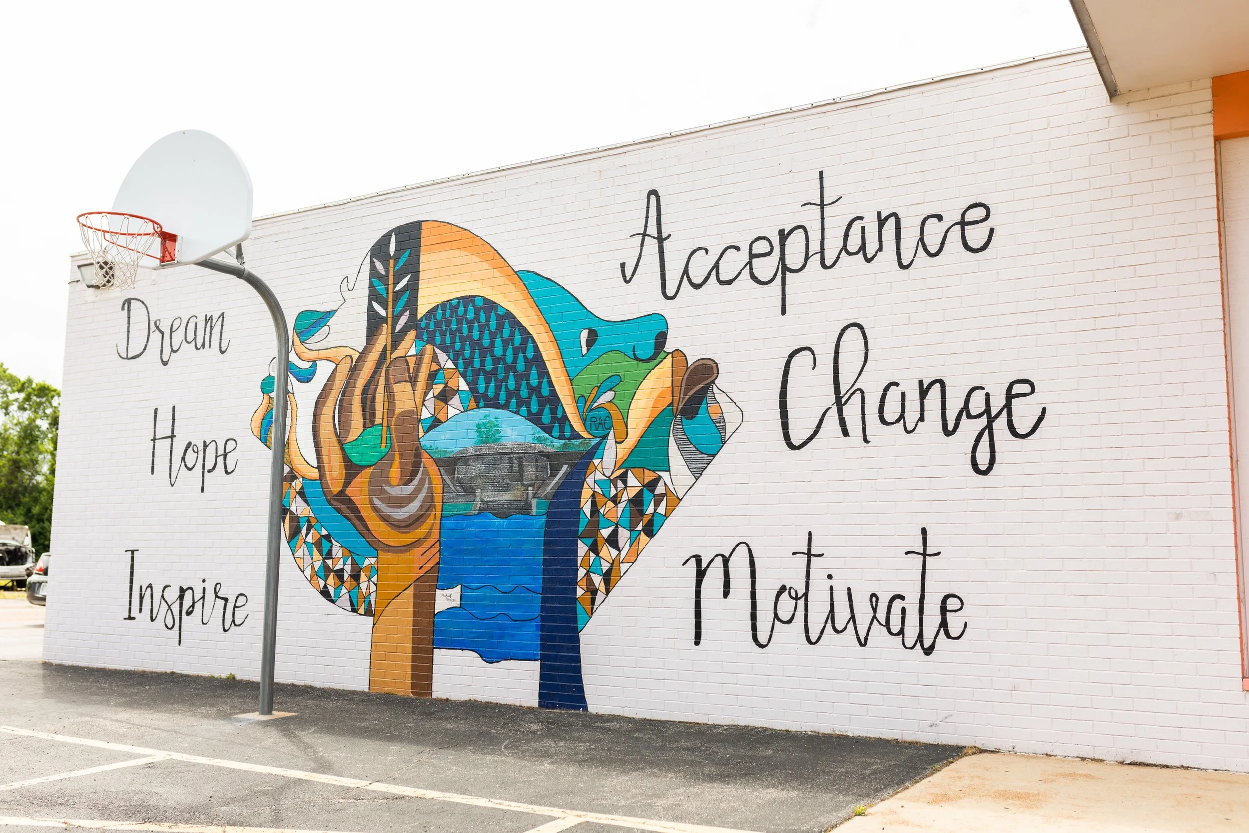 Colorful mural on a white brick wall featuring a tree, blue water, and a landscape with the words "Dream Hope Inspire" on the left and "Acceptance Change Motivate" on the right, with a basketball hoop in front.