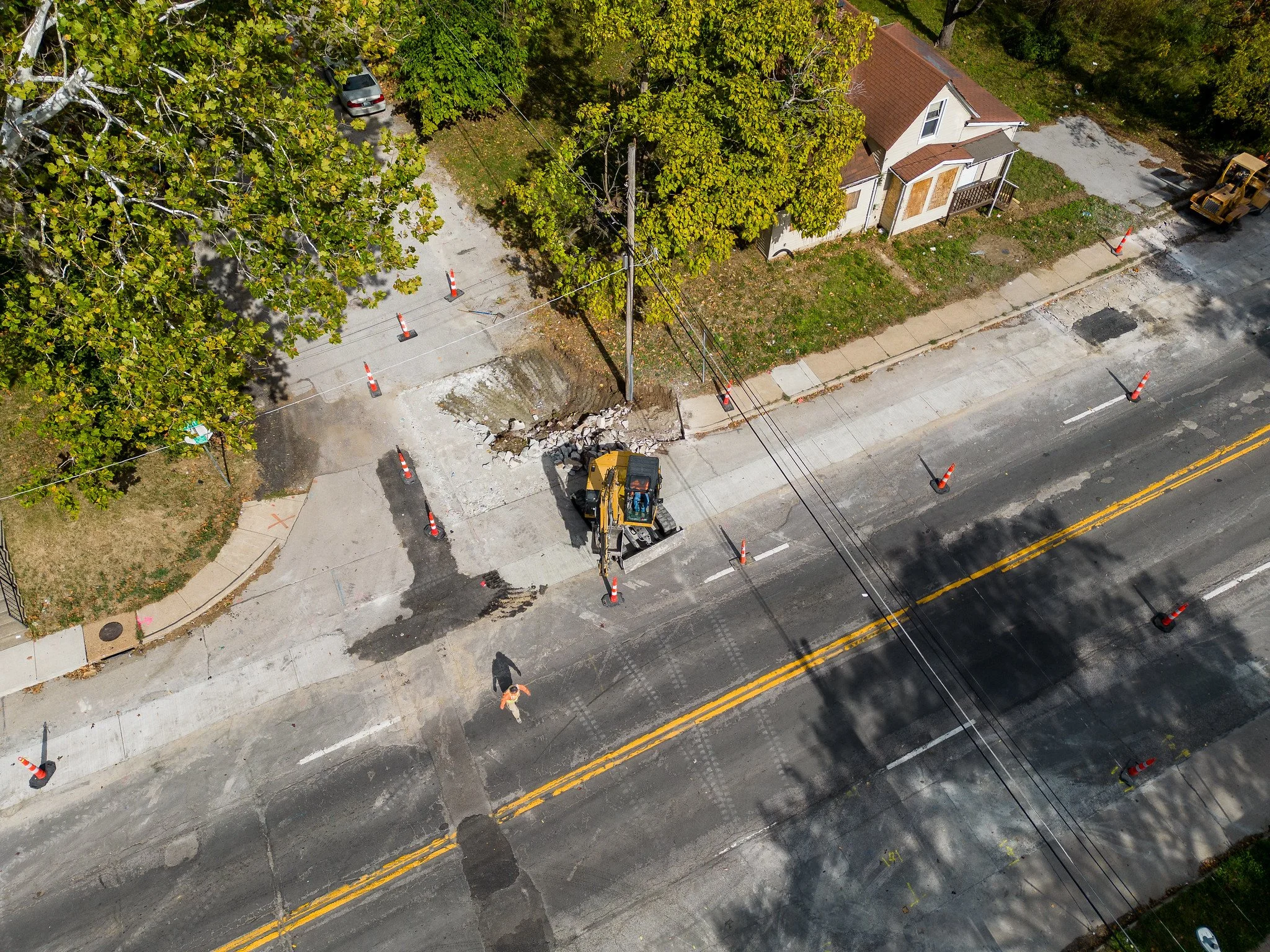 An overhead view of a road construction site with orange traffic cones placed around a section of the sidewalk and street, a small excavator working on the sidewalk, and a person walking nearby. There are trees, a house, and parked cars visible.