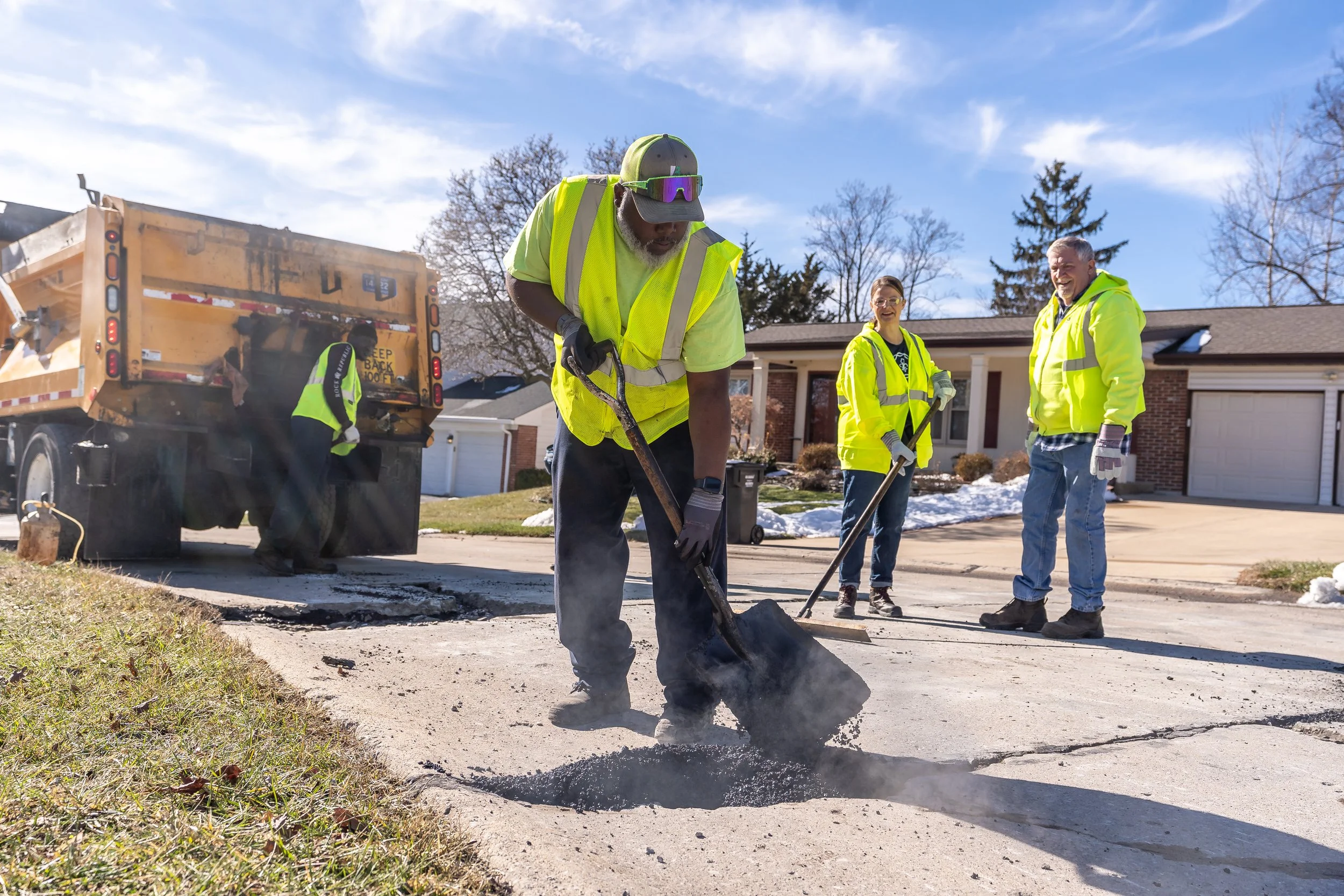 Workers in bright yellow safety vests and jackets repairing a residential street, with a team of four people, asphalt patching, and a dump truck on a sunny day.
