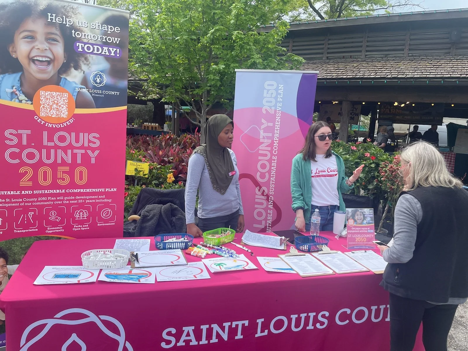 Community outreach booth set up with pink tablecloth promoting Saint Louis County 2050 plan. Two women, one wearing a hijab and the other with sunglasses, are talking to a woman with gray hair. The booth displays flyers, pens, and a small water bottle, with banners in the background highlighting the plan's goals for development, transportation, economic opportunity, parks, open space, and environment.