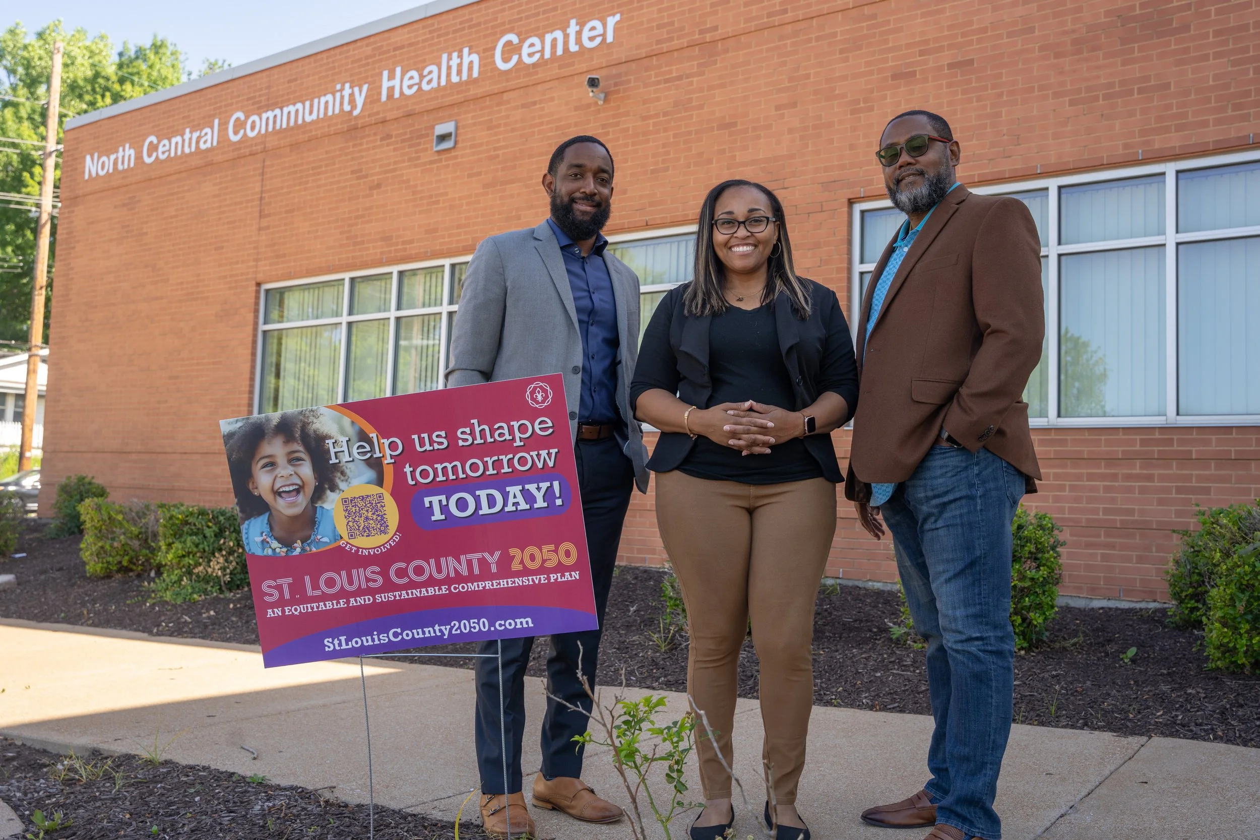 Three people standing outside the North Central Community Health Center, holding a sign promoting a plan for St. Louis County 2050. The sign features a smiling child's picture and a QR code, calling for community involvement.