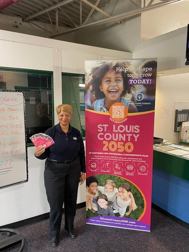 A woman standing indoors next to a promotional banner for St. Louis County 2050, holding pink hand fans with the same logo. The banner features smiling children and text about shaping the future of St. Louis County.