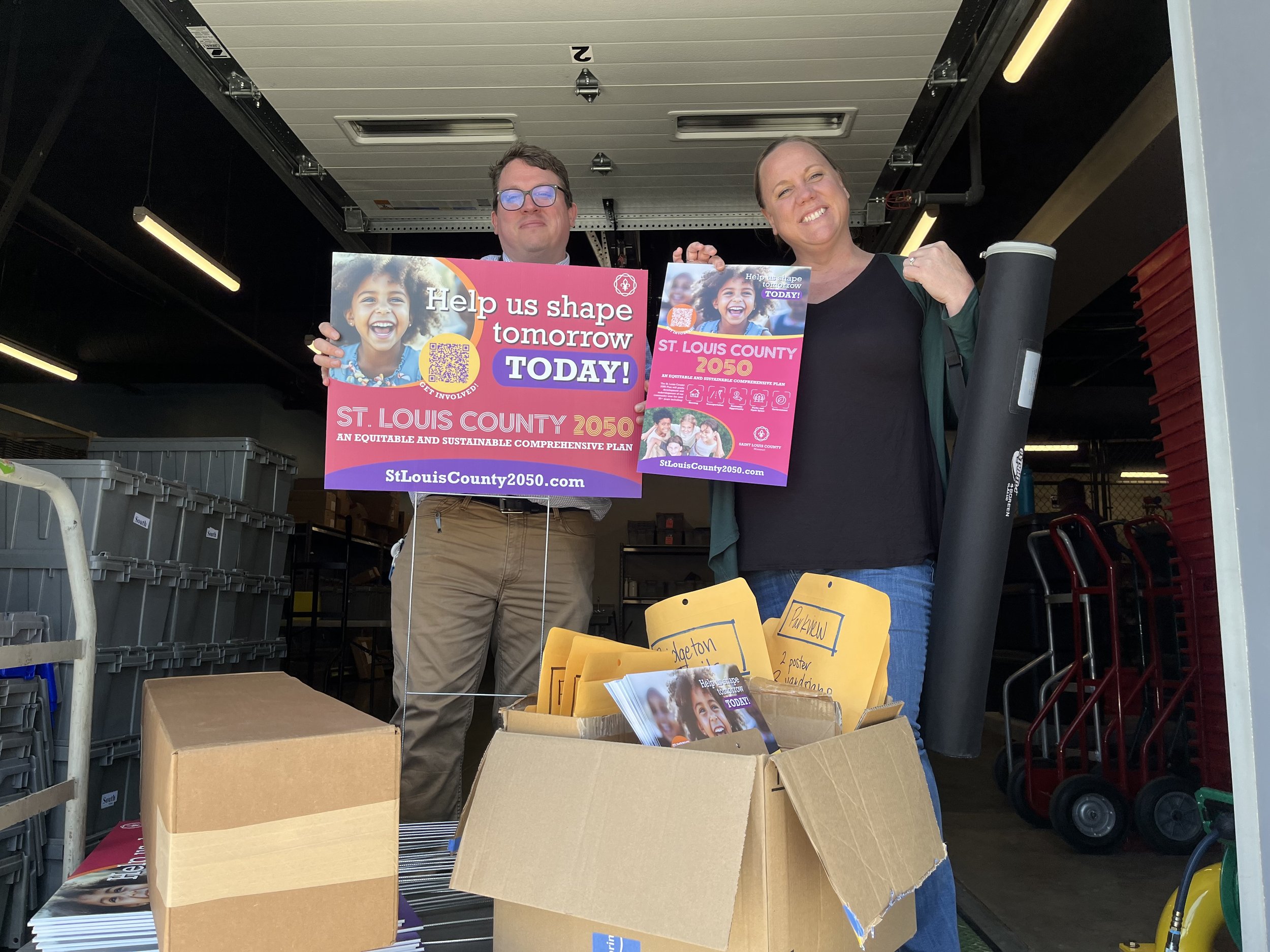 Two people standing inside a truck holding signs supporting St. Louis County 2050 plan. One woman is holding a rolled-up poster, and there is an open cardboard box with flyers and envelopes in the foreground.