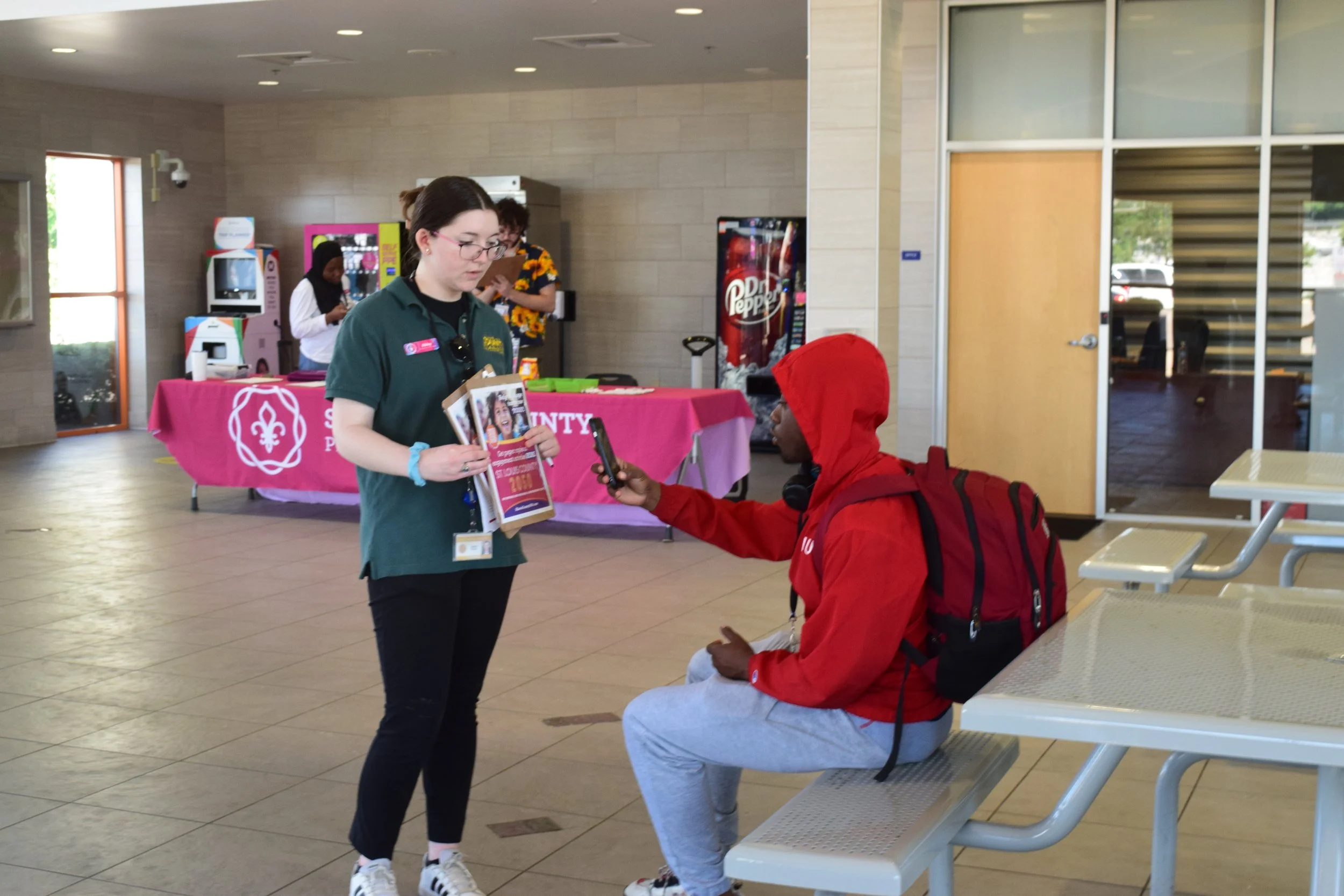 A woman in a green uniform and a man in gray sweatpants sitting on a bench, both holding phones, inside a building with a table in the background covered with a pink cloth, vending machines, and a door.