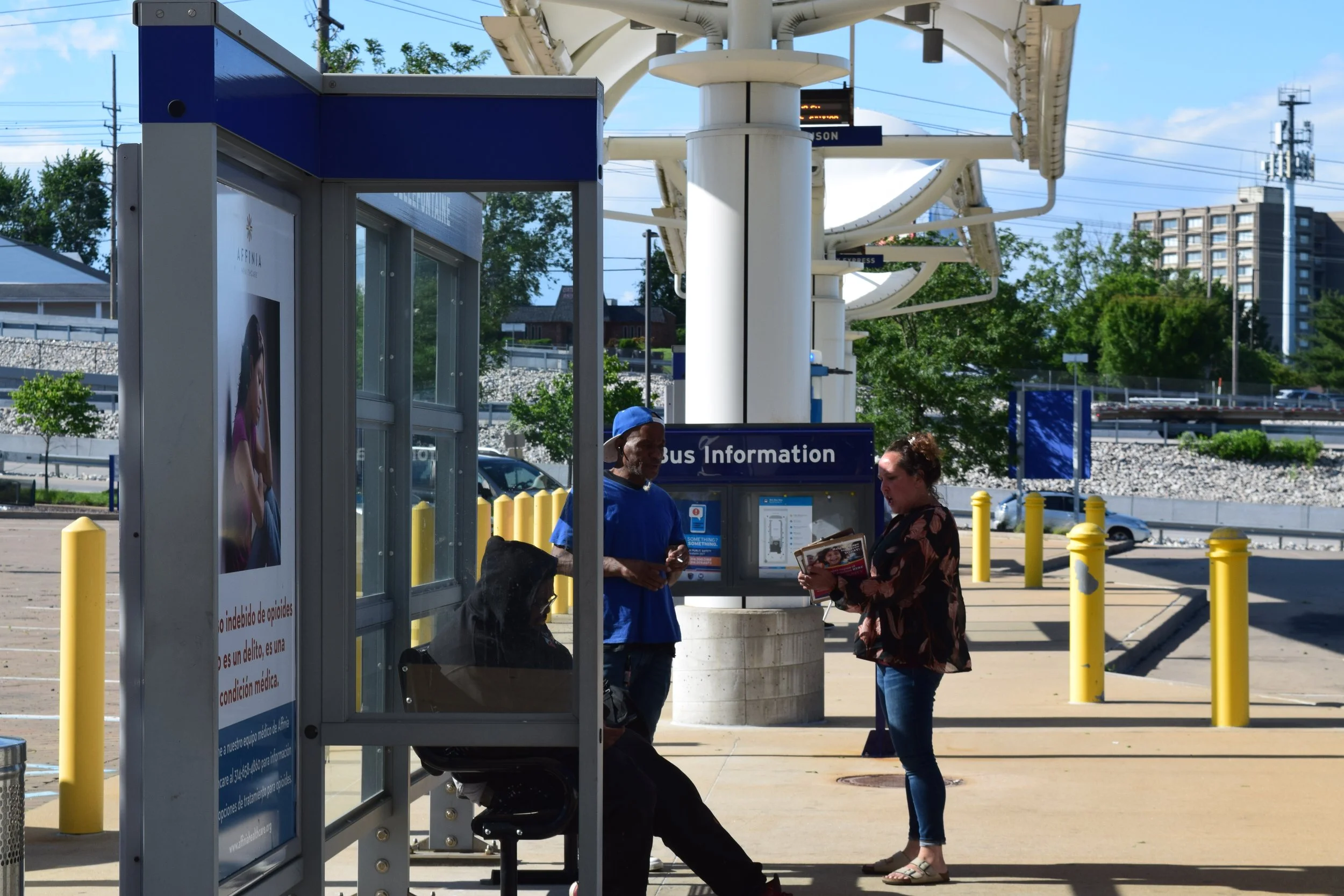 People waiting at a bus stop with bus information sign, shelter, and advertisements, near a city street with trees and buildings in the background.