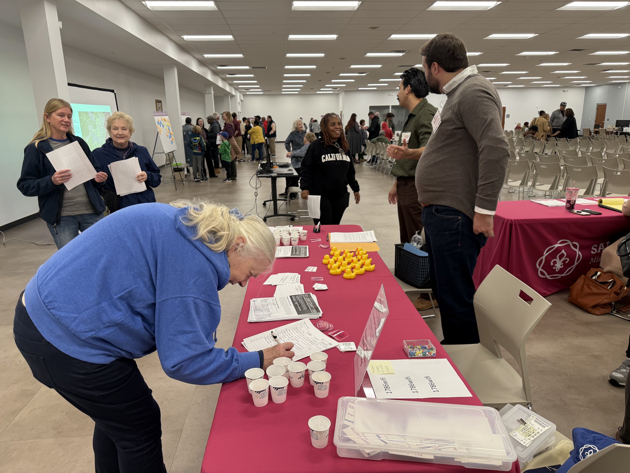 People attending an event at a large indoor hall with tables and chairs. Several individuals are standing and talking near a table with informational materials, rubber duckies, and cups. In the background, more people are gathered, some looking at displays or tables.