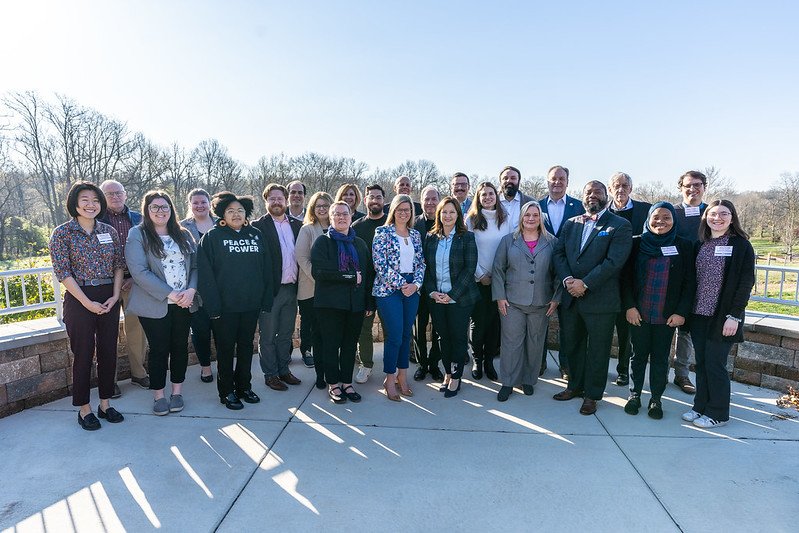 Group of diverse people standing outdoors on a sunny day, posing for a photo, with trees in the background and shadows on the ground.