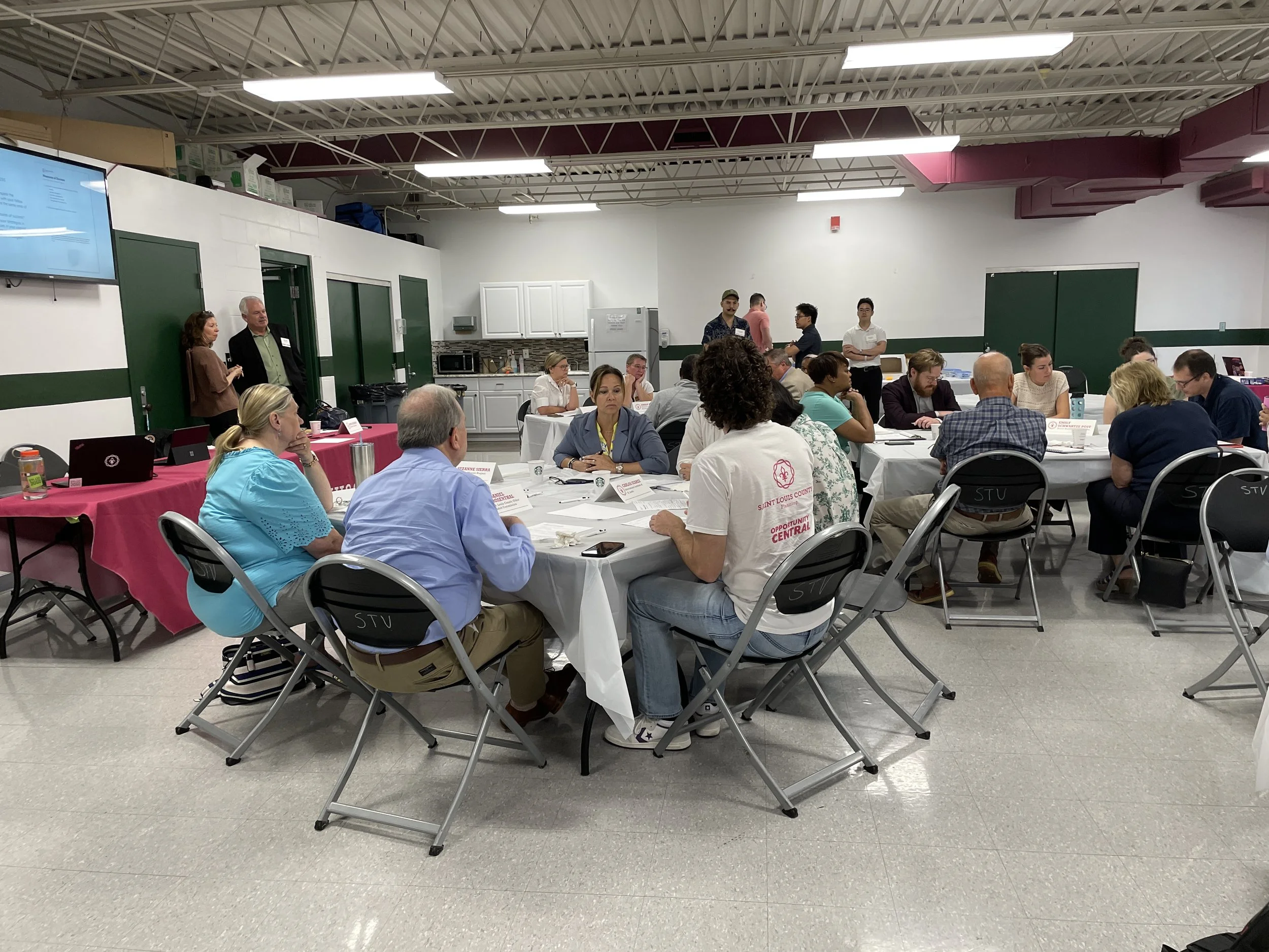 People sitting at round tables participating in a meeting or conference inside a large room with white walls and green accents.