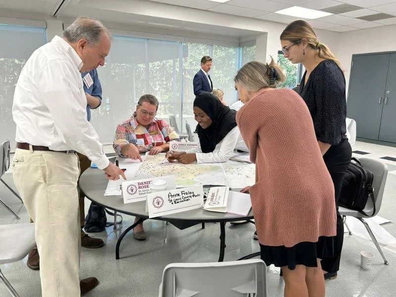 Group of diverse people gathered around a table, looking at maps during a meeting or workshop, with name tags and papers on the table in a bright room with large windows.