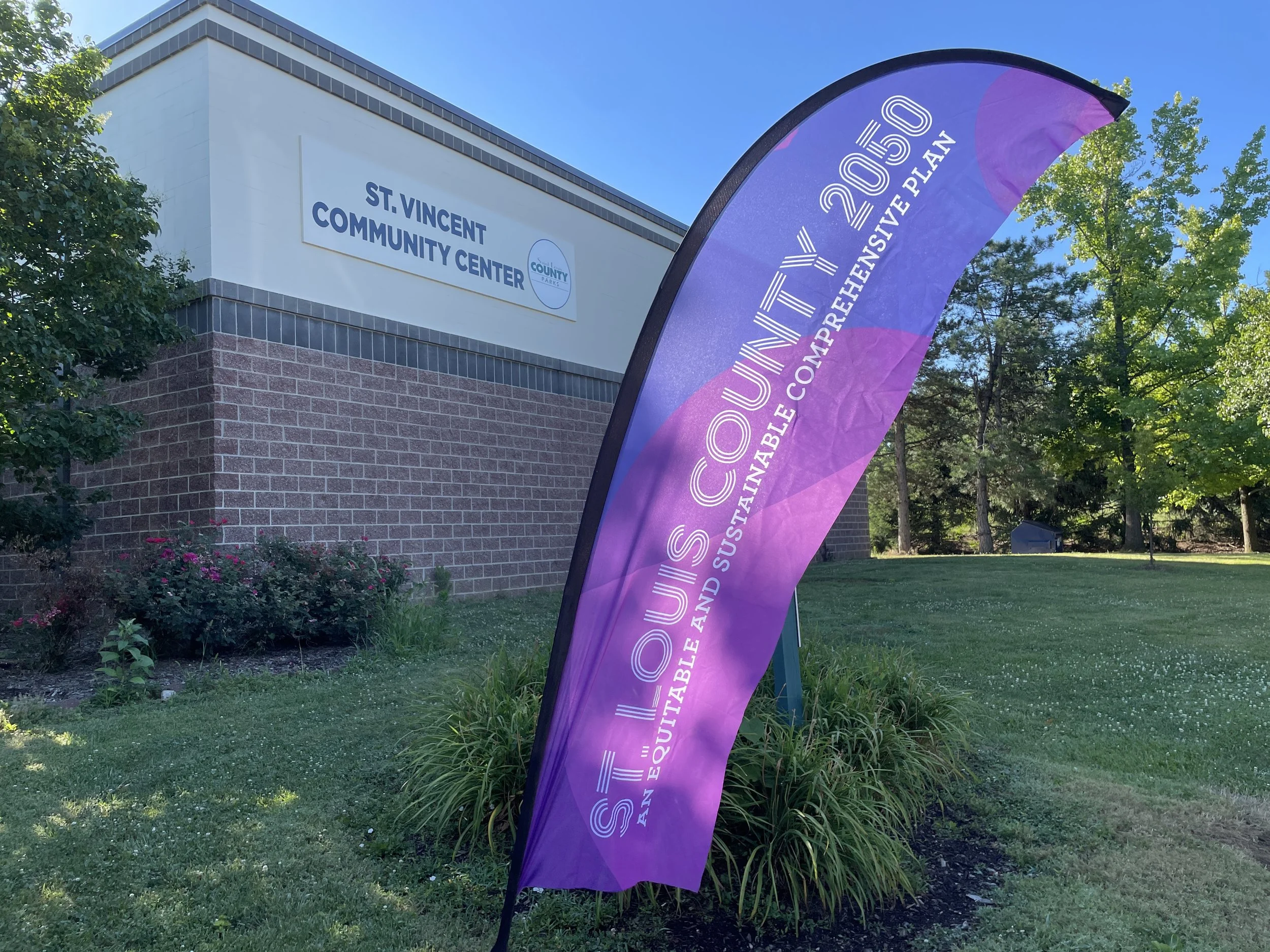 A purple and pink teardrop-shaped banner with the text 'SUSTAIN AND INNOVATE 2030 AN EQUITABLE AND SUSTAINABLE COMMUNITY' in front of the St. Vincent Community Center building, which has a brick base and a white upper section with a sign reading 'ST.