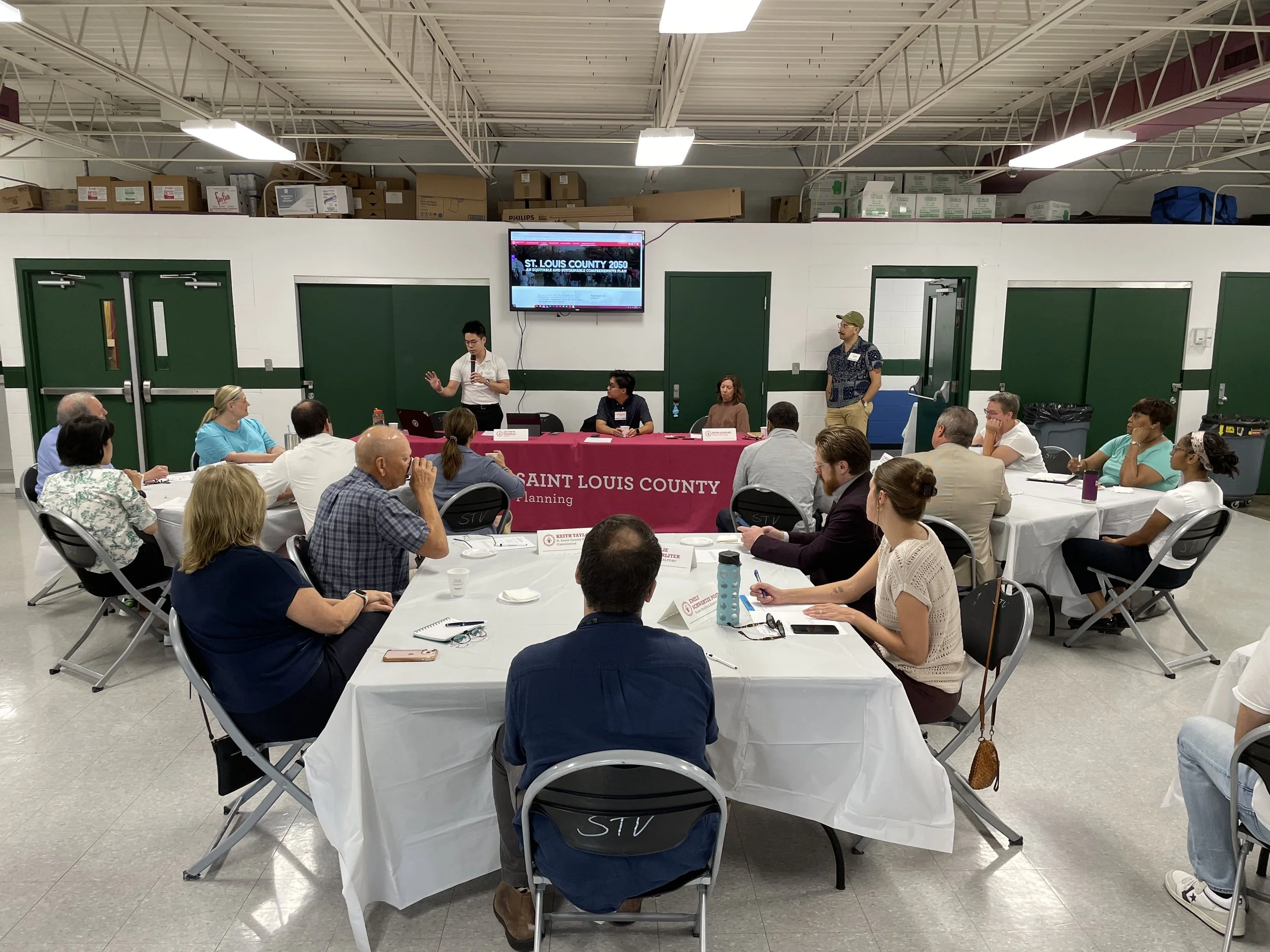 A meeting or panel discussion with multiple people seated around tables, some with laptops and notes, in a large room with industrial ceilings, a large screen on the wall displaying "ST. LOUIS COUNTY 2050," and a table in front labeled "SAINT LOUIS C
