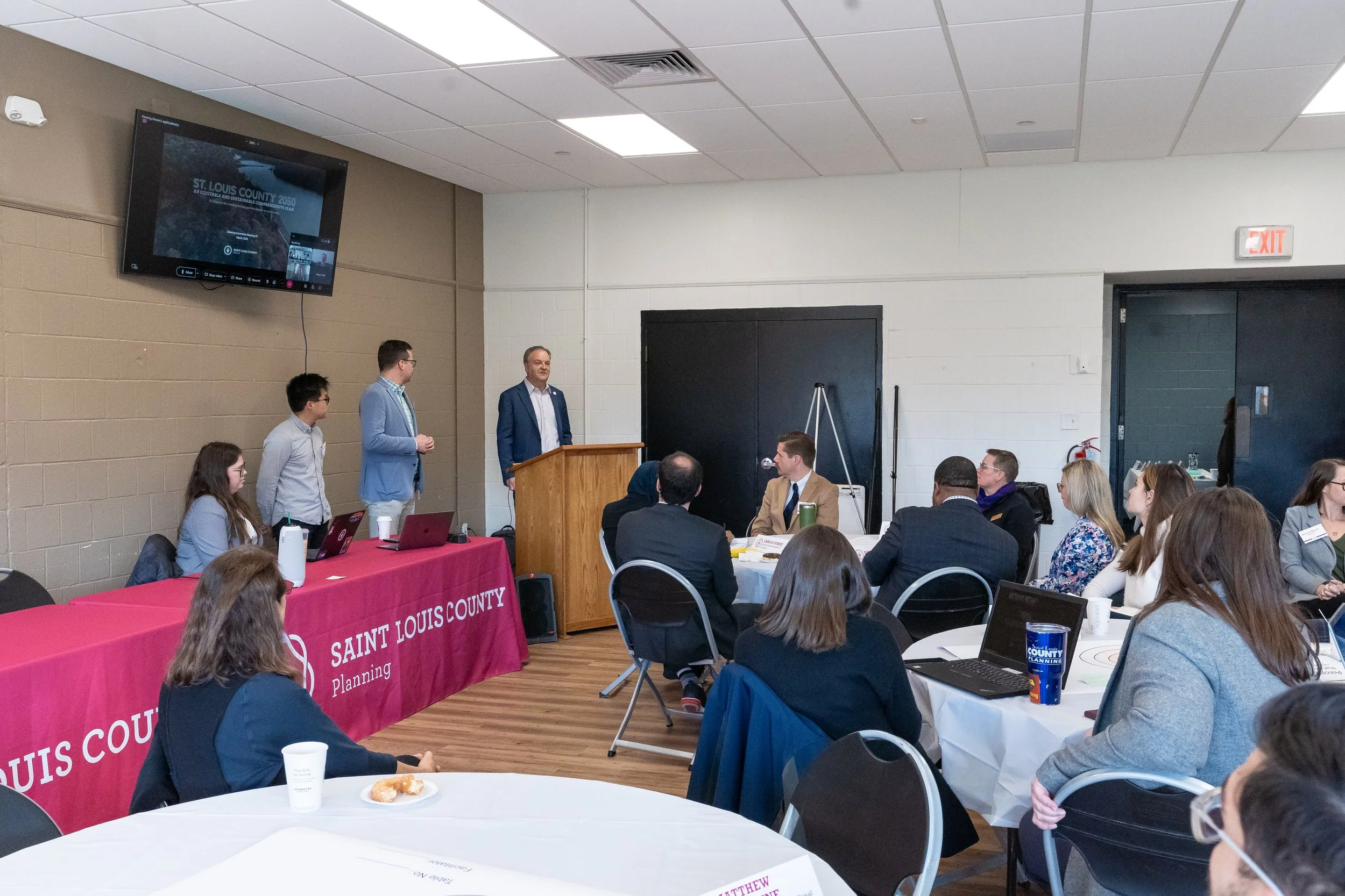 A group of people attending a conference or meeting in a room with a man speaking at a wooden podium. A large screen showing the presentation and a pink tablecloth with 'Saint Louis County Planning' printed on it are visible.