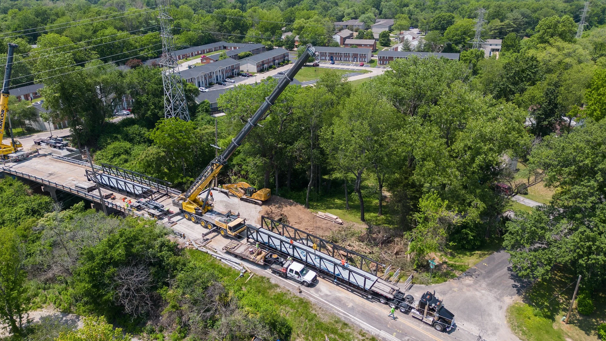 A construction site on a bridge with a crane lifting a steel beam, surrounded by trees and residential houses in the background.
