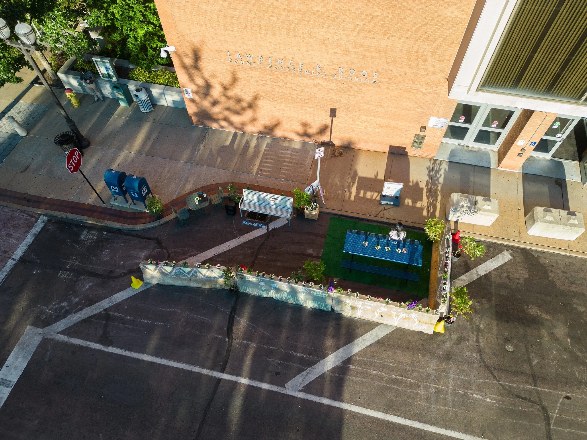 Aerial view of downtown street scene featuring a small fenced area with a ping pong table, benches, potted plants, and flowers, adjacent to a brick government building with the name "Lawrence K. Roos County Government Building."