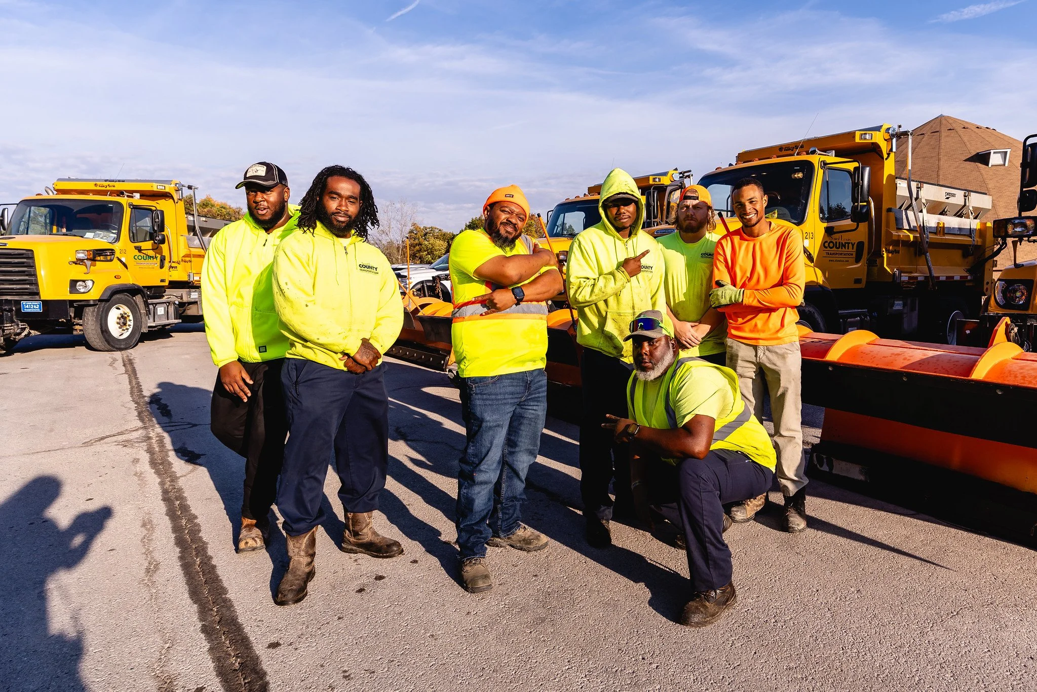 Group of workers in high-visibility clothing standing in front of snowplows and yellow trucks during daytime.