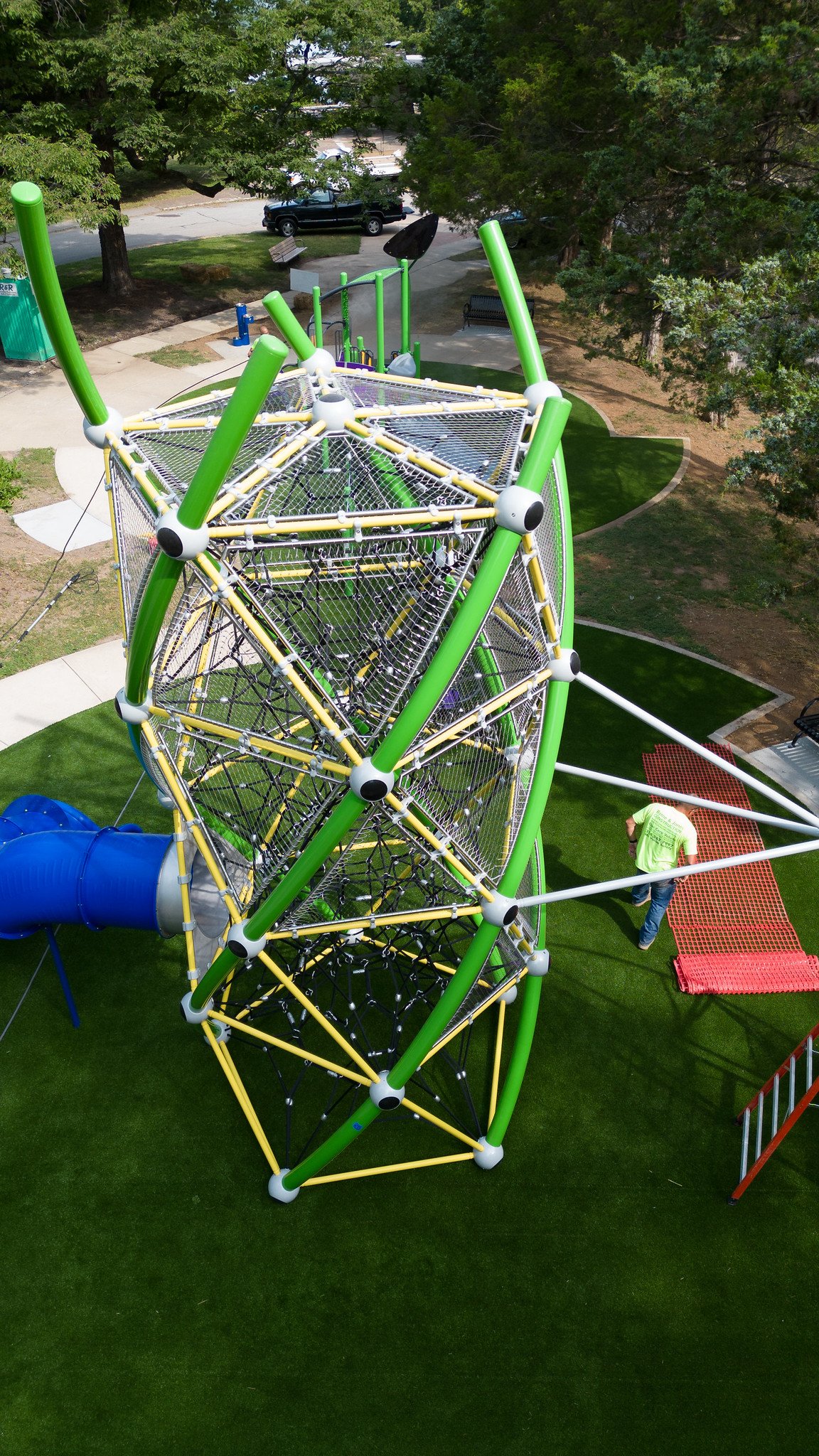 Colorful outdoor playground structure with climbing nets and a blue slide, set on artificial turf, surrounded by trees and benches.