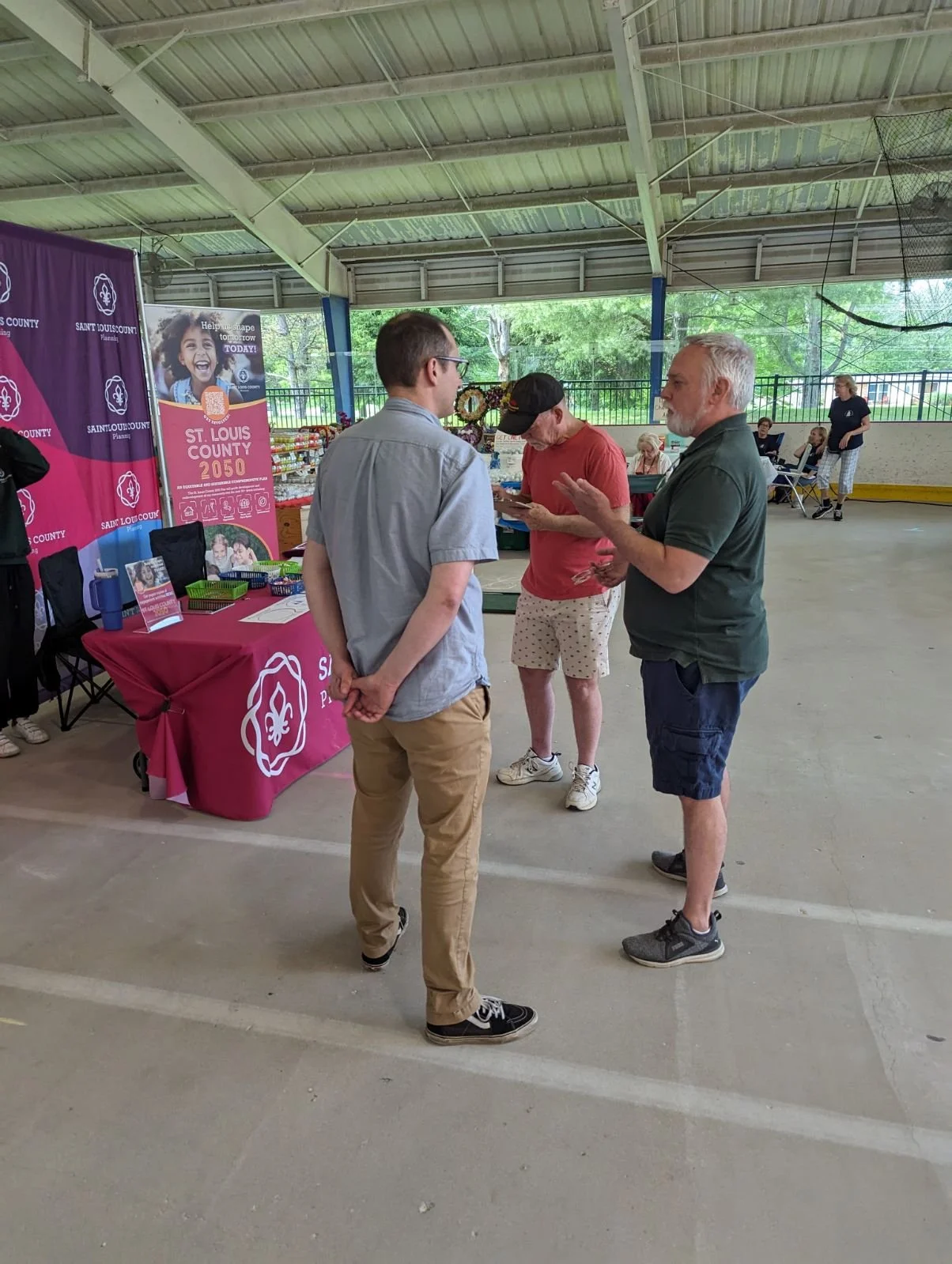 People talking at a Saint Louis County booth inside a pavilion with a purple banner and a pink tablecloth.