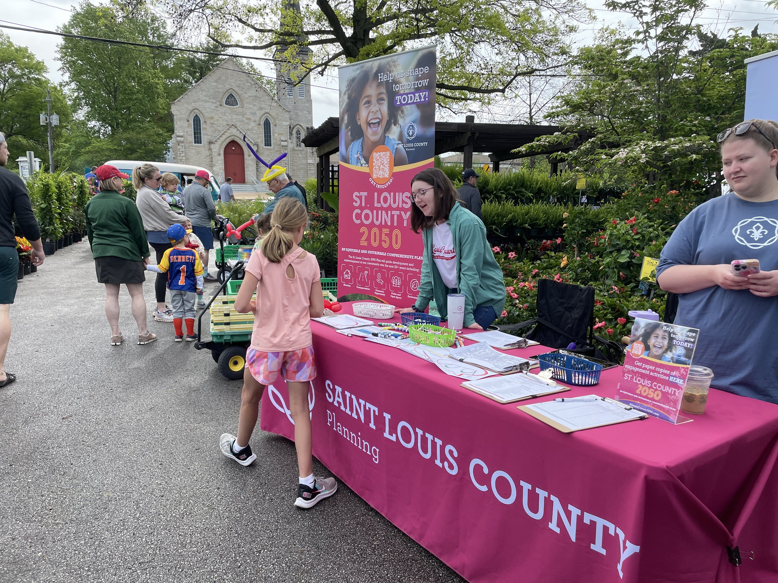 Children and adults at a community event, with a table for Saint Louis County planning displaying informational materials and a banner promoting future planning goals. The event takes place outdoors near a church and greenery.