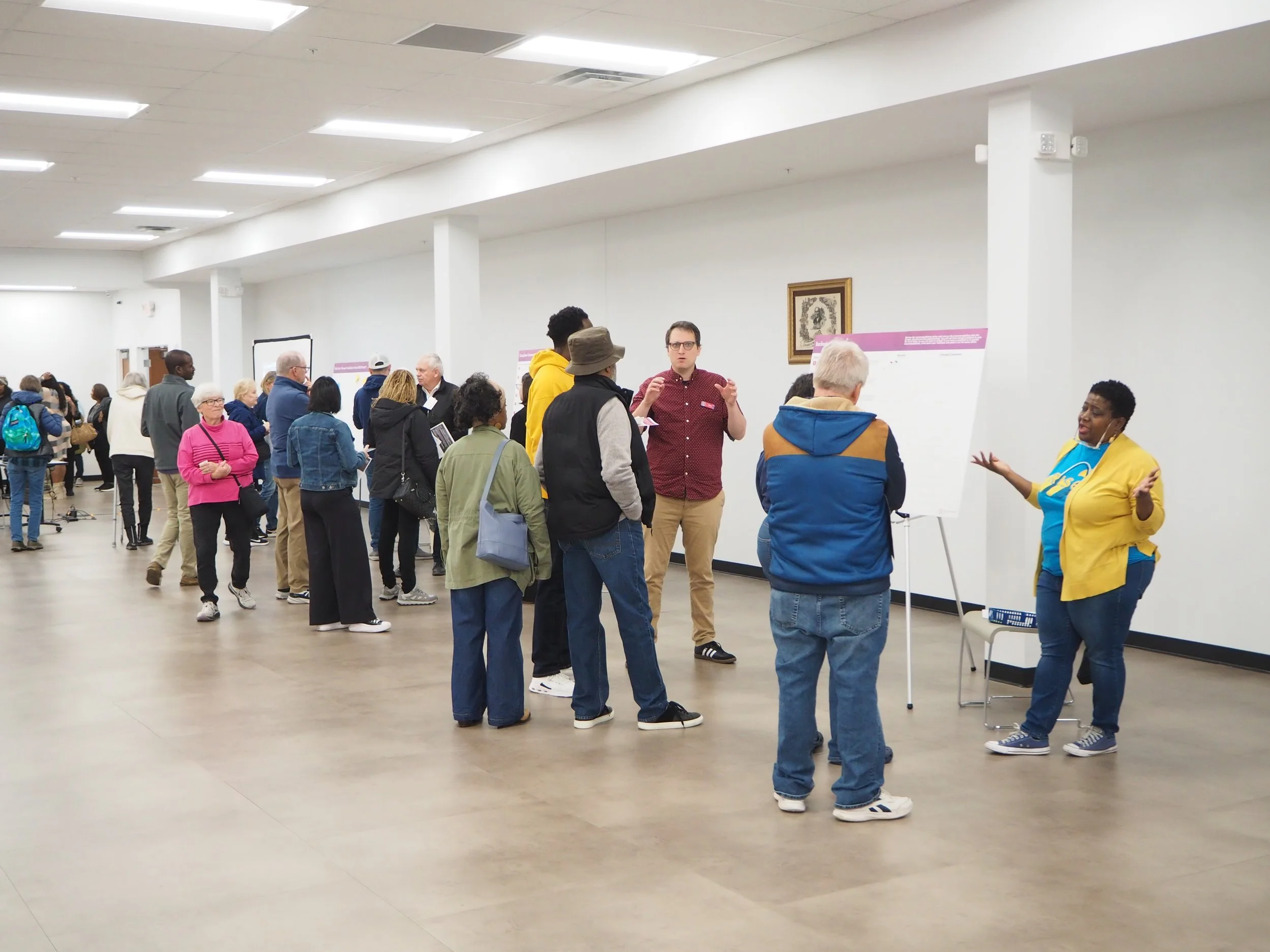 People attending a seminar or workshop in a large room, with one person speaking to the group and others standing in line or listening.