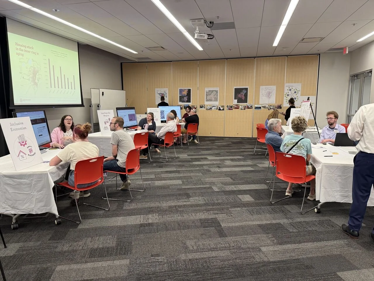 Conference room with tables and red chairs where people are engaged in discussions and presentations, with posters and maps displayed on the wall.