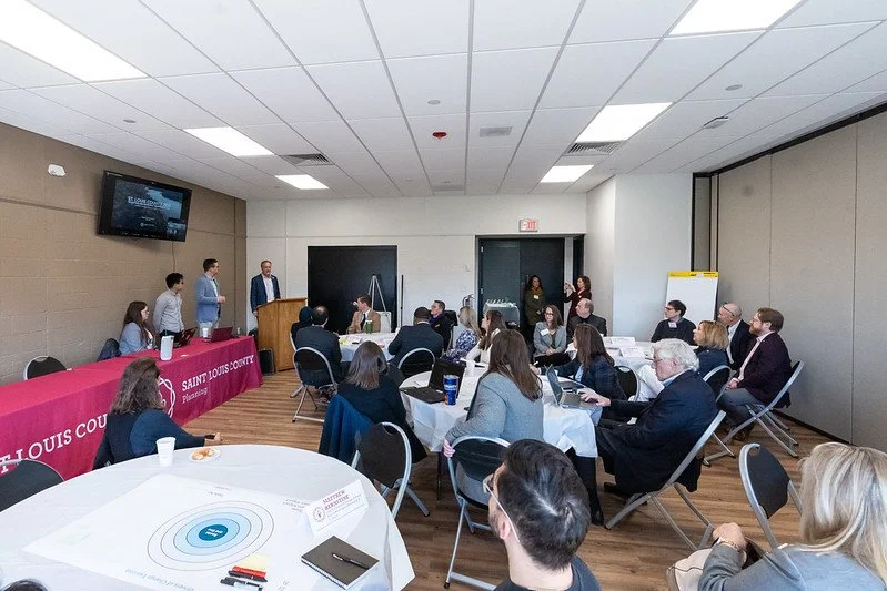 A group of people attending a meeting or presentation in a conference room with tables, chairs, and a large screen on the wall.