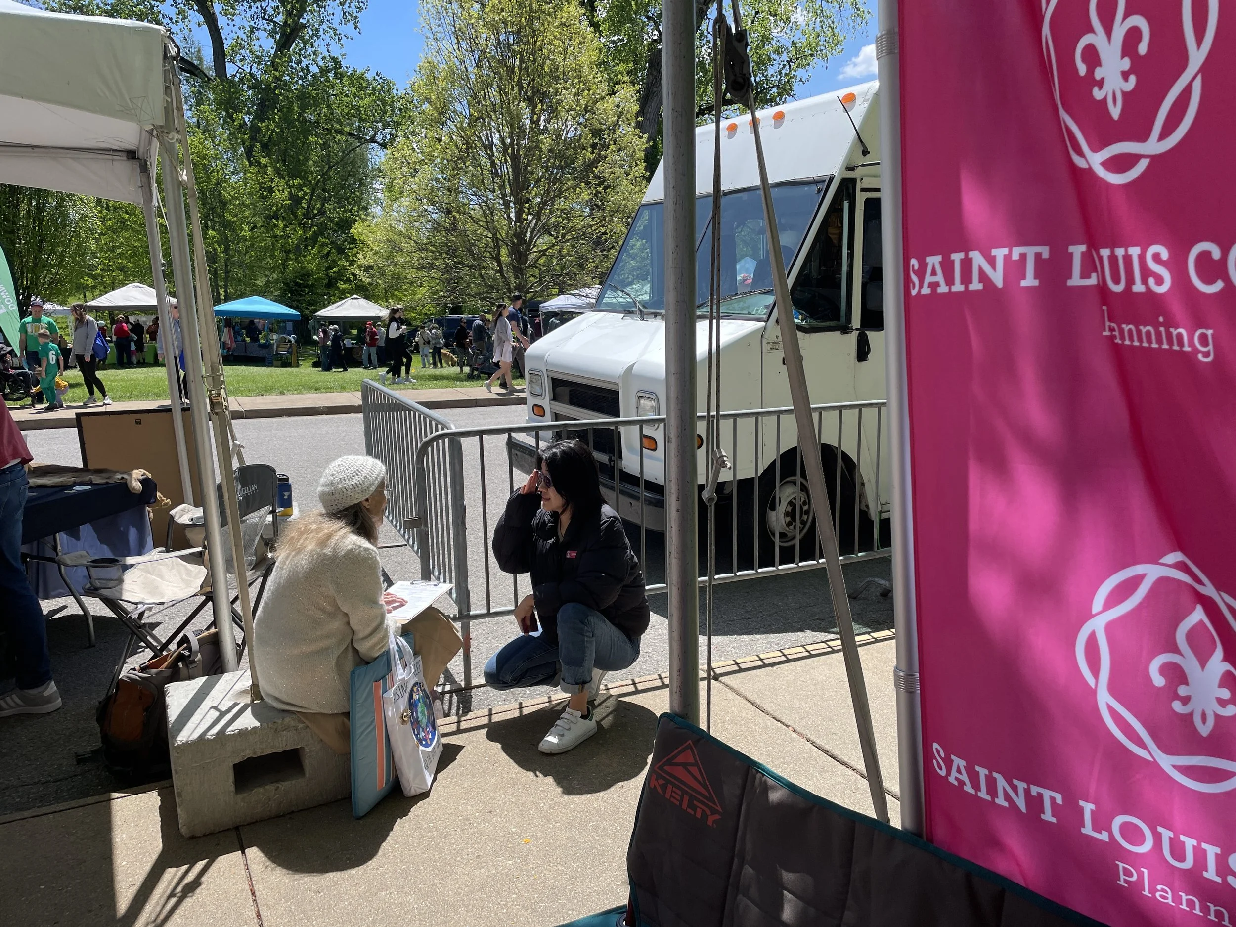 Two women seated and talking at an outdoor event booth on a sunny day, with tents, people, and trees in the background. One woman has long gray hair and a white knit cap, the other has dark hair and sunglasses. A pink banner with white text reads "SAINT LOUIS CO" and "anning."