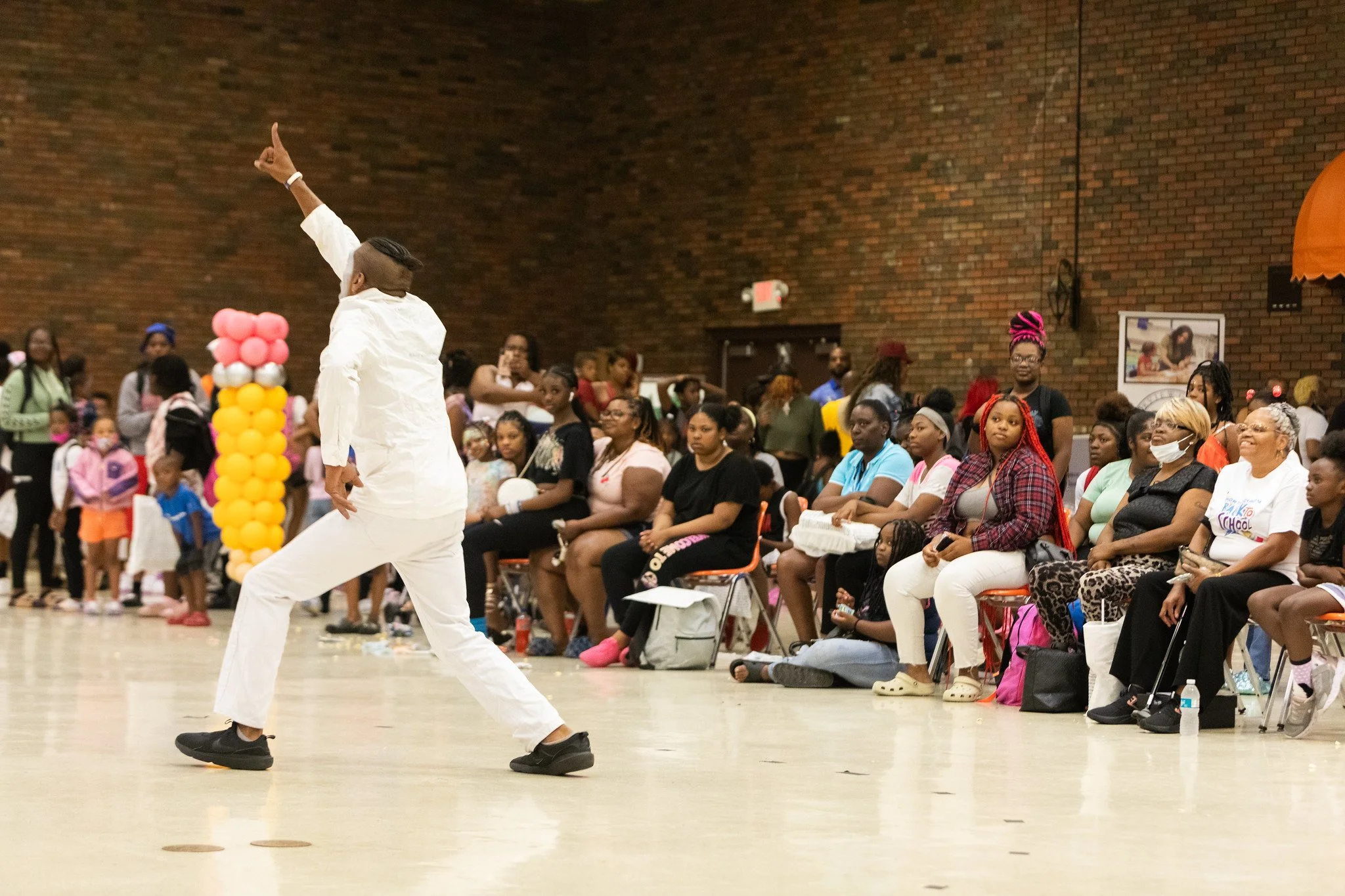 A performer in white pants and shirt, with a mohawk hairstyle, dancing on stage in front of an audience. The audience, consisting mostly of women and children, are seated and standing against a brick wall in the background, watching and enjoying the performance. There are colorful balloons arranged in a column on the stage.