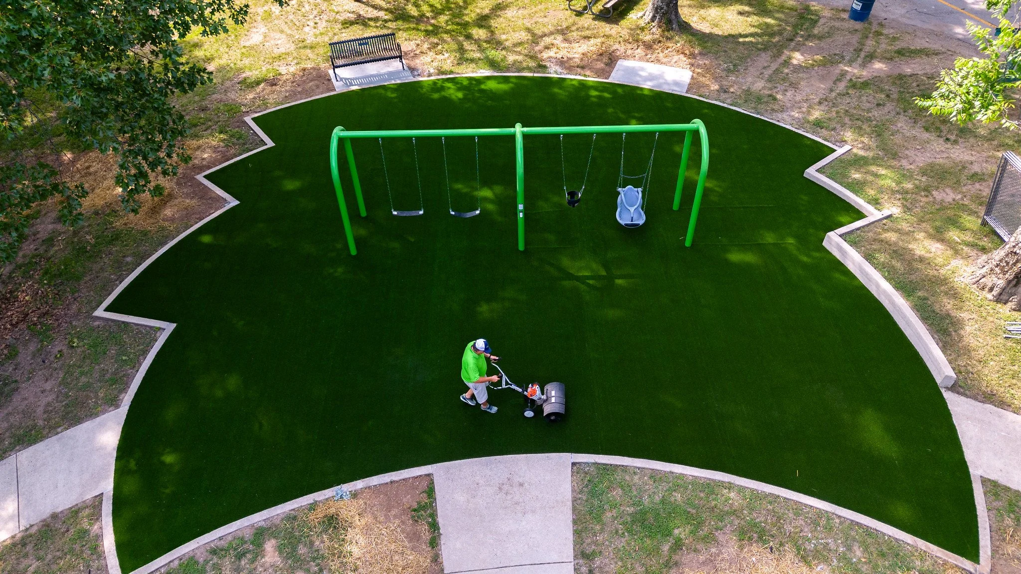 An aerial view of a park with a green artificial turf area, a swing set with four swings, including a baby swing, and a person mowing the grass with a walk-behind reel mower. There are benches, trees, and a sidewalk surrounding the turf area.