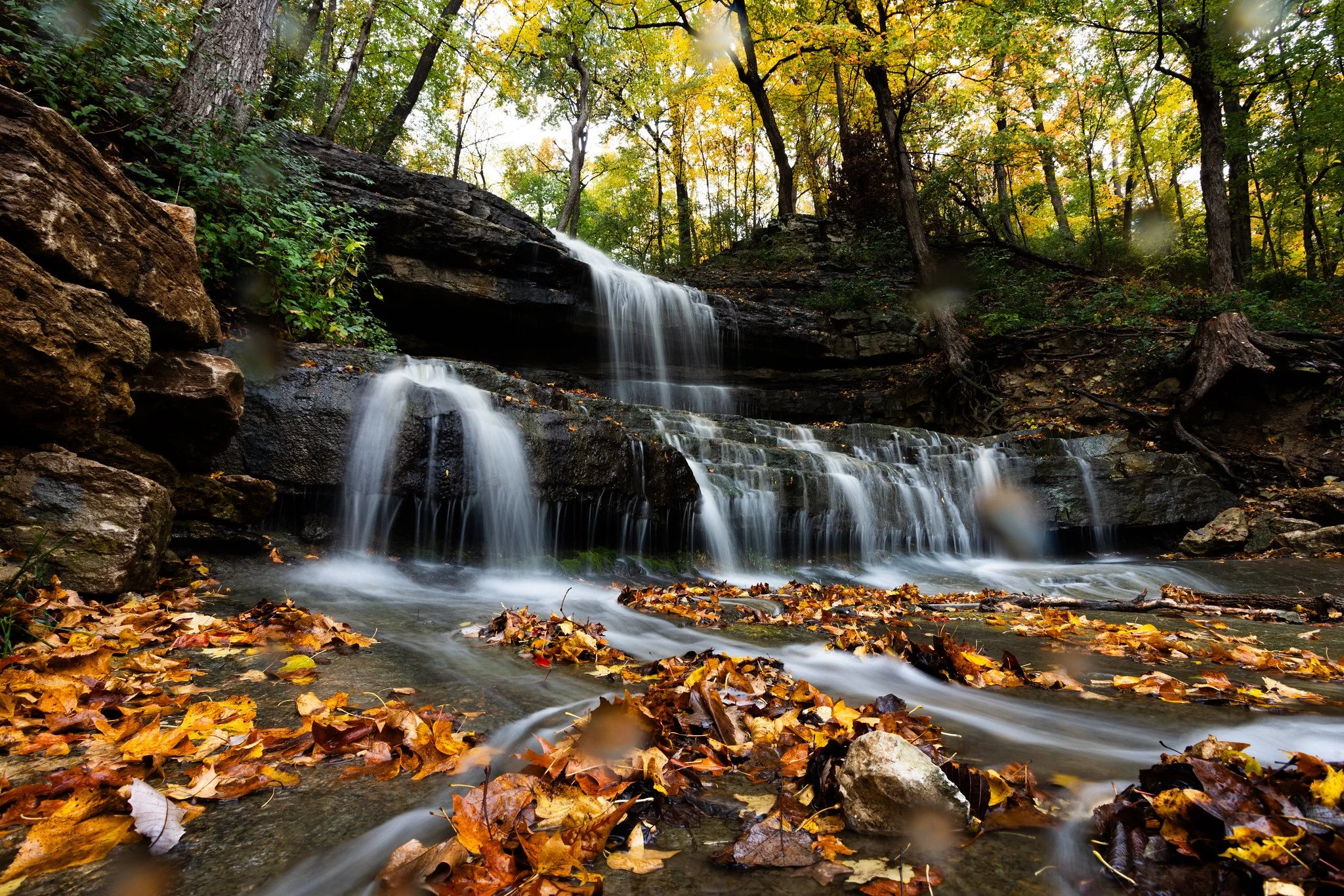 A multi-tiered waterfall flowing over rocks in a forest with colorful autumn leaves on the ground and trees with green, yellow, and orange foliage.