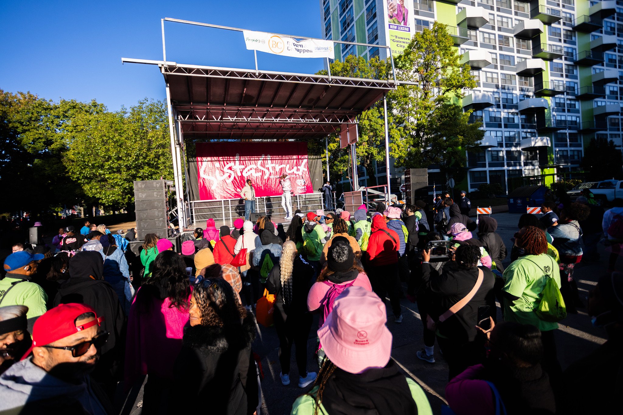 Crowd of people attending an outdoor event or rally with a stage, some wearing colorful hats and jackets, in front of a modern residential building with trees and blue sky.