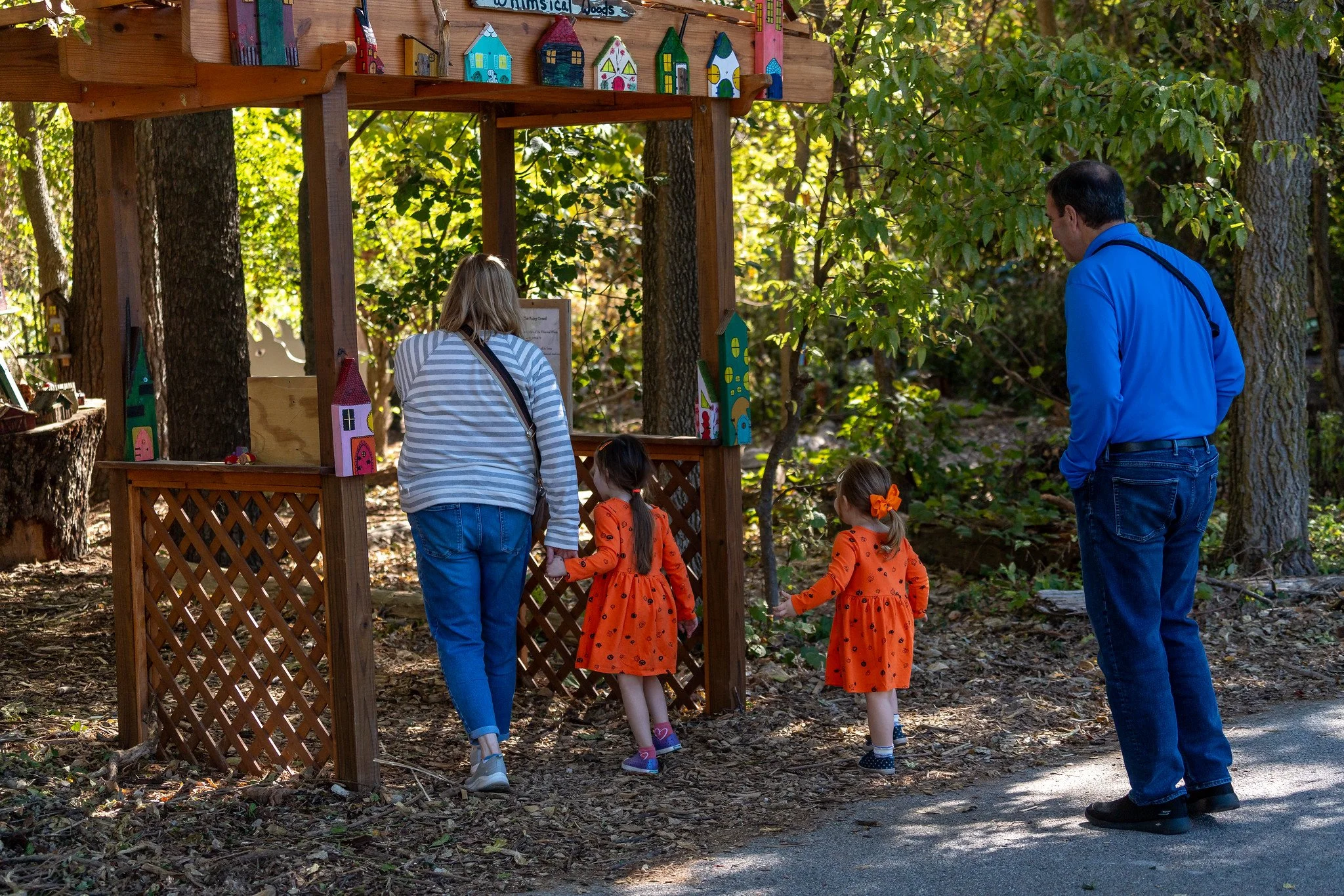 A family of four, two adults and two young girls, observe birdhouses displayed on a wooden stand in a wooded area.