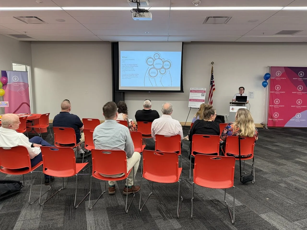 A small conference room with attendees sitting on red chairs, facing a presentation screen. A woman stands at a podium on the right side, speaking. There are banners on the sides with the Saint Louis County logo, balloons, and an American flag.