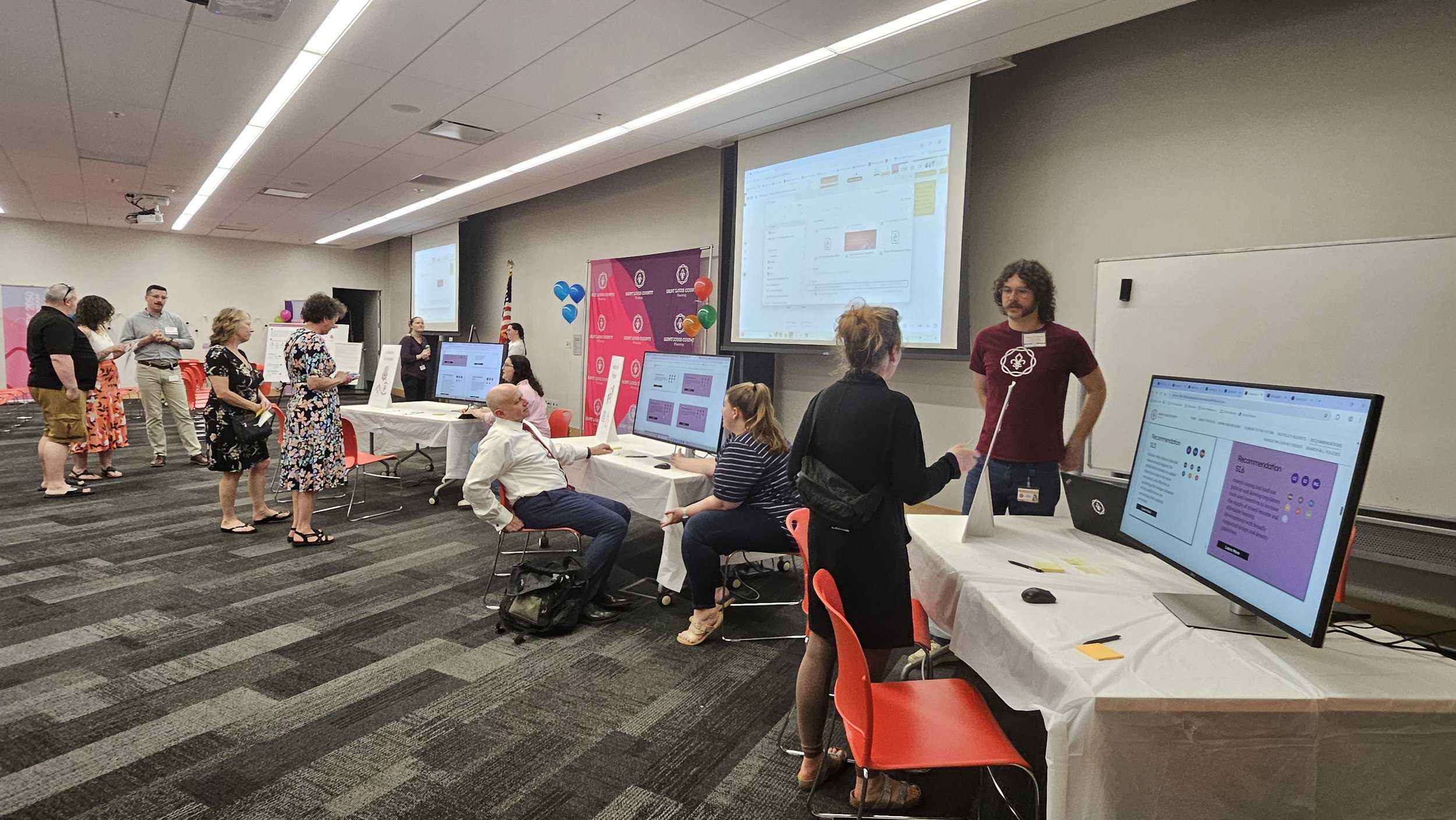 People in a conference room participating in a registration or check-in process, with multiple tables, computer monitors, and large screens displaying information.