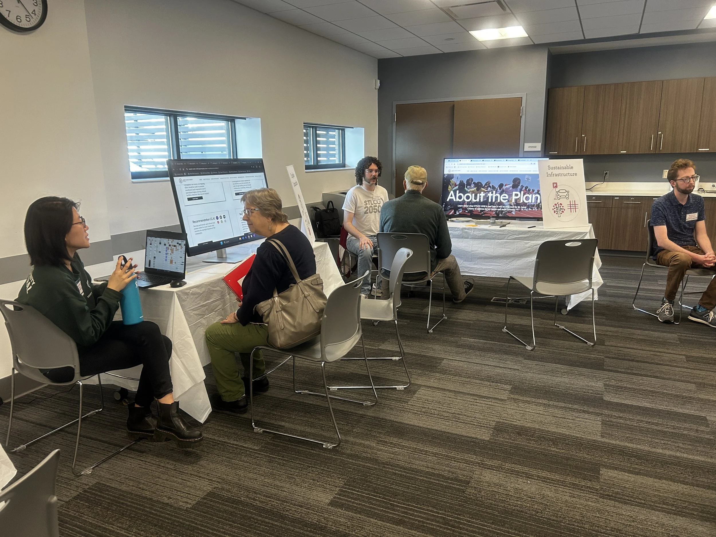 A conference room with multiple people sitting at tables with laptops and monitors, engaged in discussions. There is a large screen with the text "About the Plan" and a sign that says "Sustainable Infrastructure." The room has windows and wooden cabi